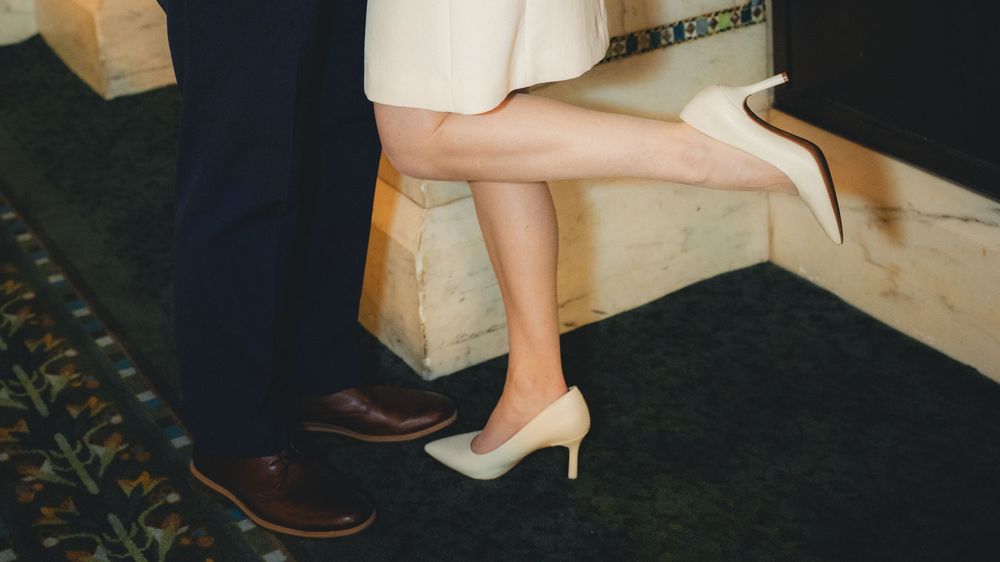 Two pairs of feet in formal shoes, one in brown dress shoes and one in beige heels, standing on wooden steps.