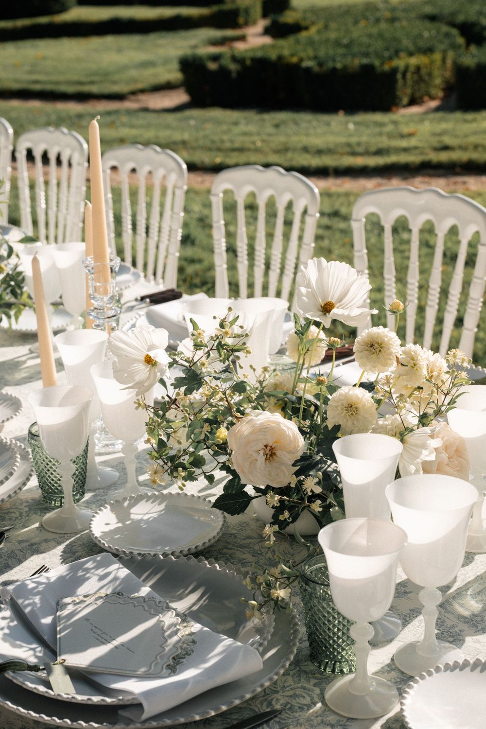 Elegant white and cream table setting with floral centerpieces and vintage-style chairs for an outdoor wedding reception.