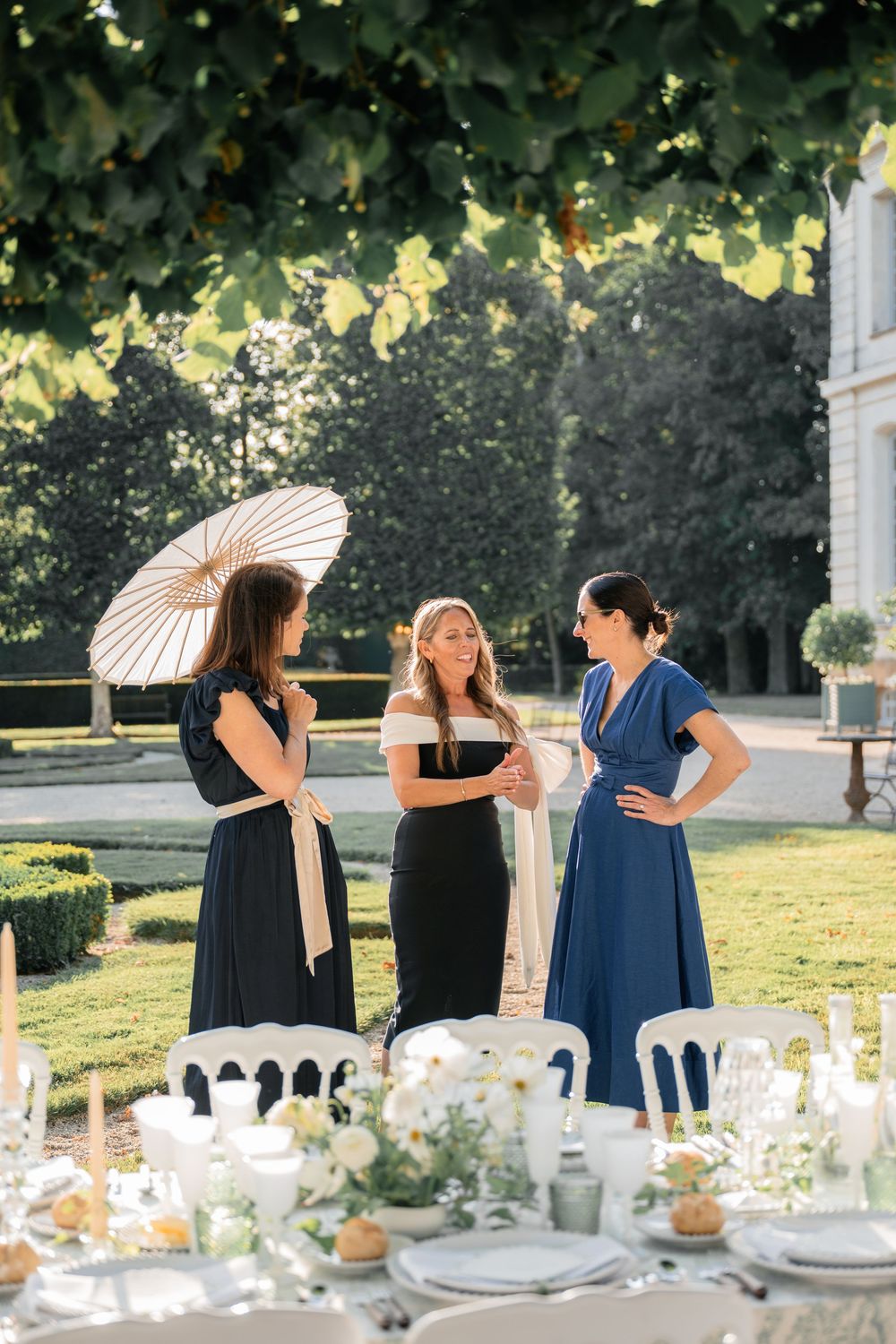 Three women in elegant dresses chat beside an outdoor wedding table setting with parasol and floral decor.
