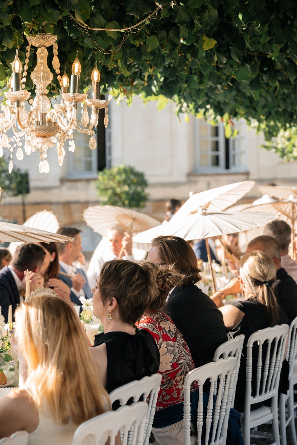 Outdoor wedding reception under trees with white chairs and crystal chandeliers creating a romantic atmosphere.