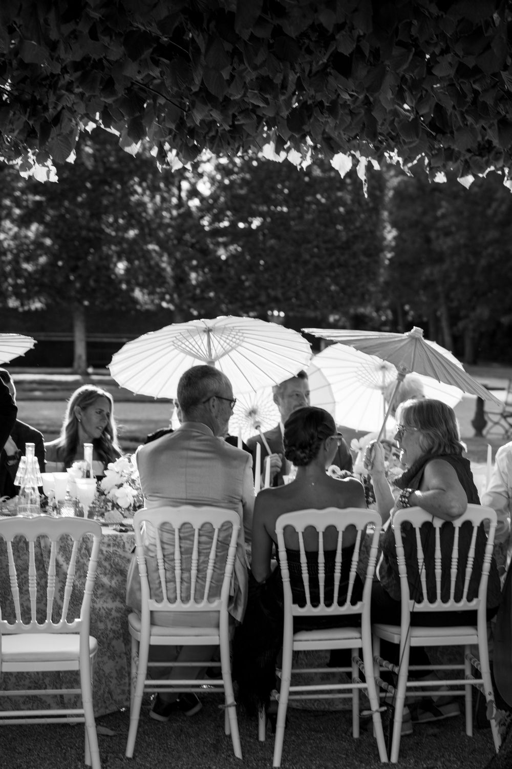 Wedding guests dine at white chairs under parasols at an elegant outdoor garden reception.