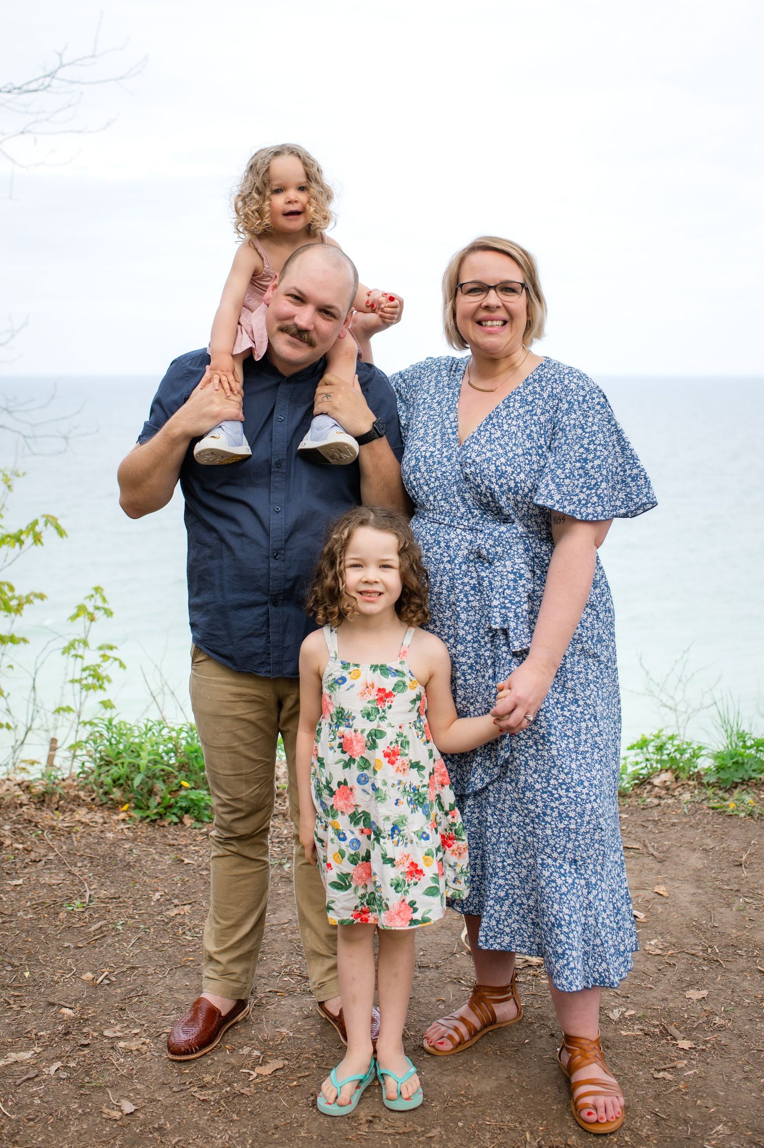 A family poses together on a cliff overlooking a lake, wearing summer attire on a bright day.