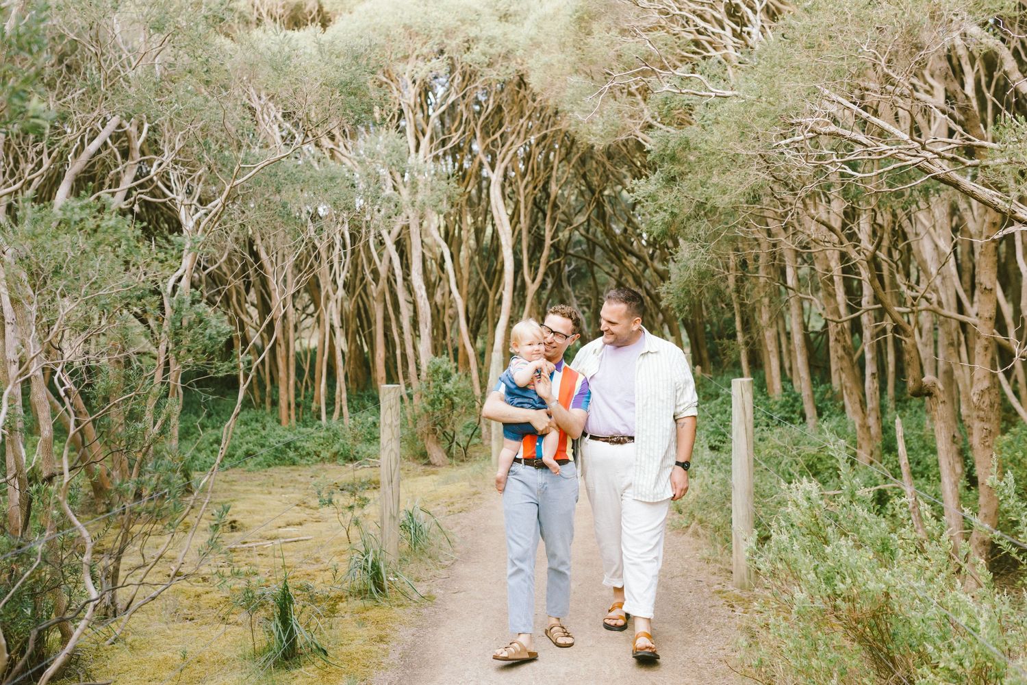 A couple walks together on a tree-lined path through a serene wooded area on a sunny day.