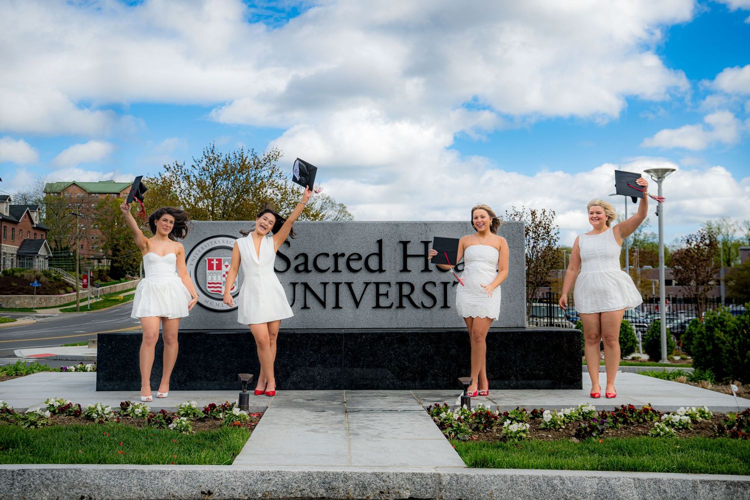 Four people wearing white dresses walk together on a sidewalk with a brick building and trees in the background.