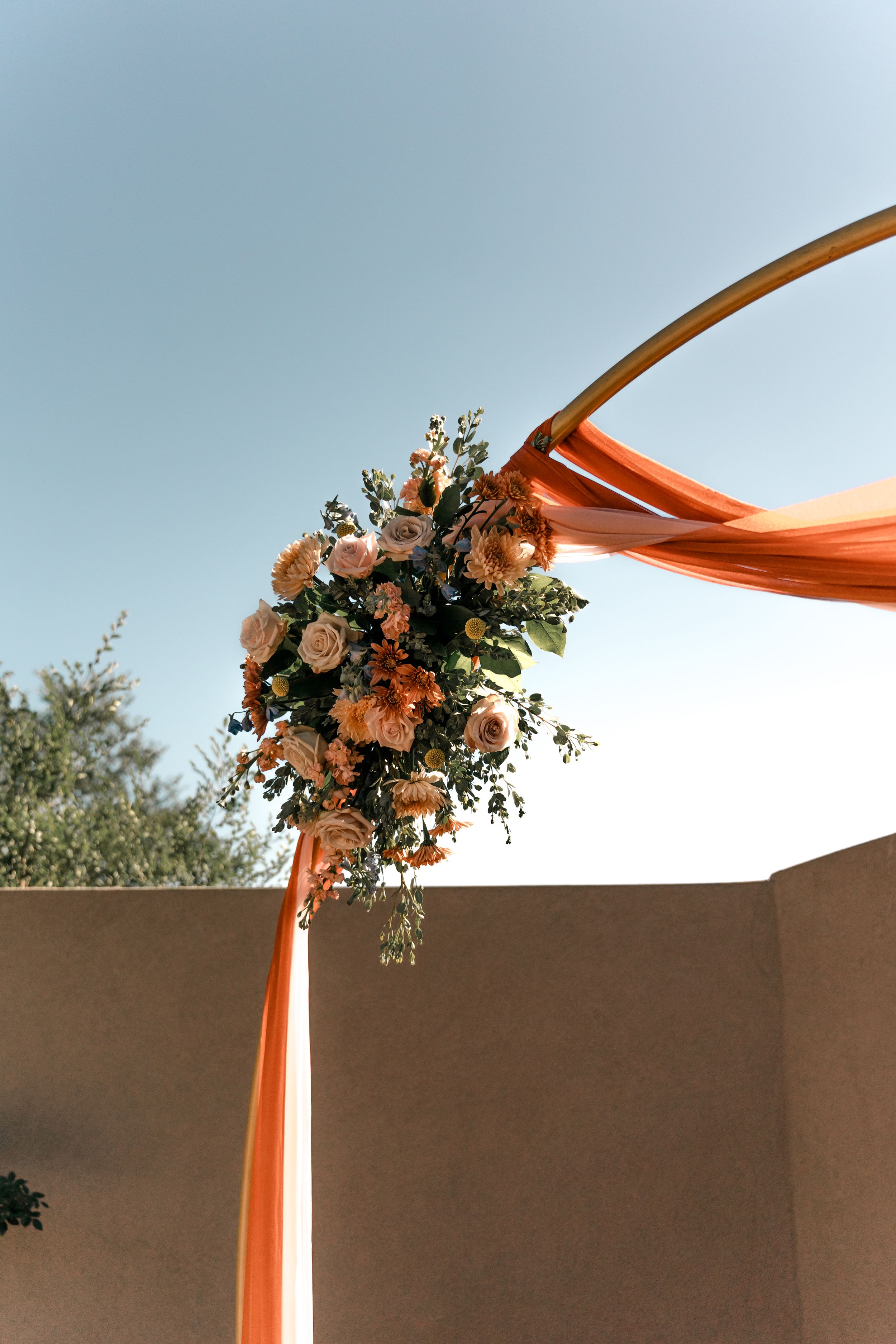 Orange fabric drapes elegantly from a wedding arch adorned with a lush floral arrangement against a beige wall.