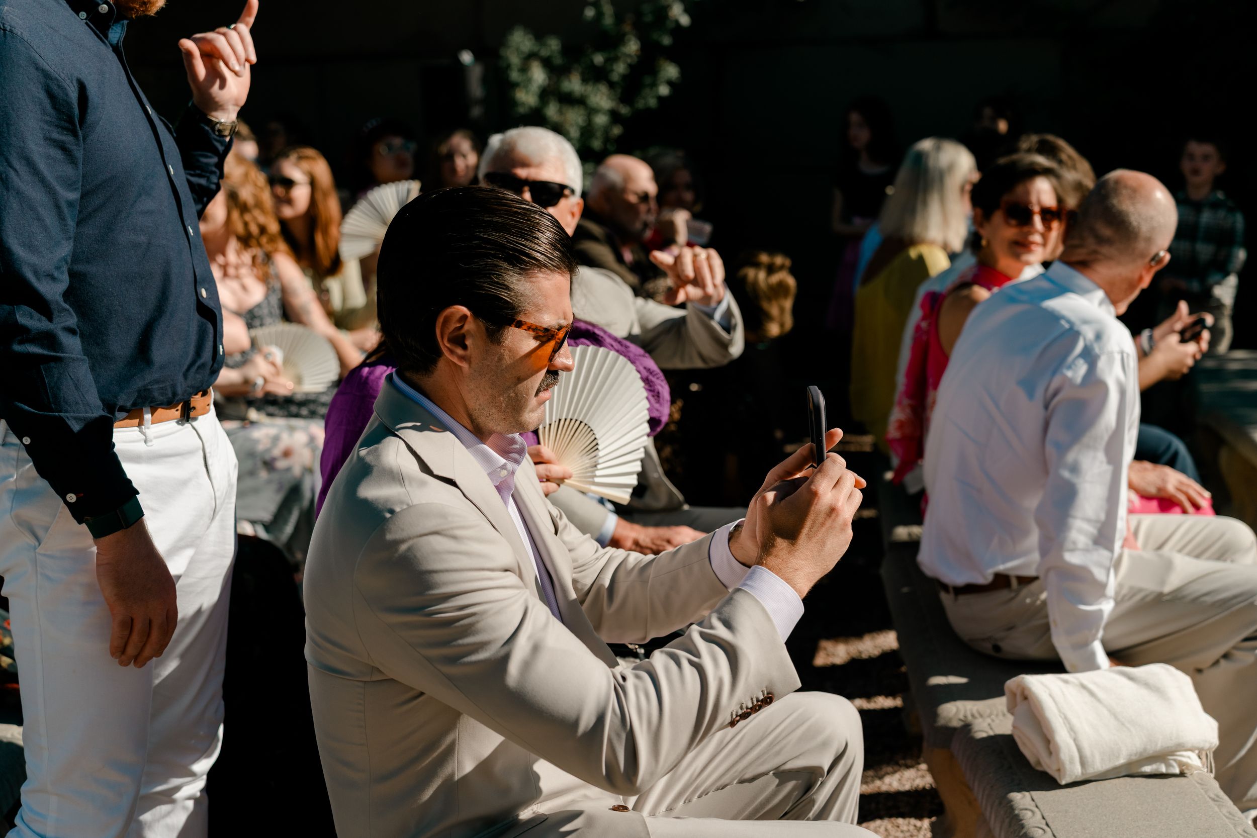 Wedding guests enjoy a sunny outdoor reception while seated at long tables under natural lighting.