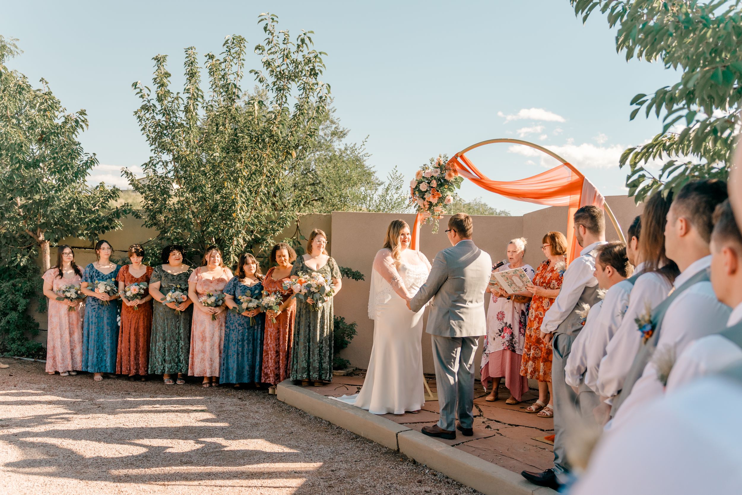 Wedding ceremony under a draped orange fabric arch with attendees gathered in formal attire outdoors.