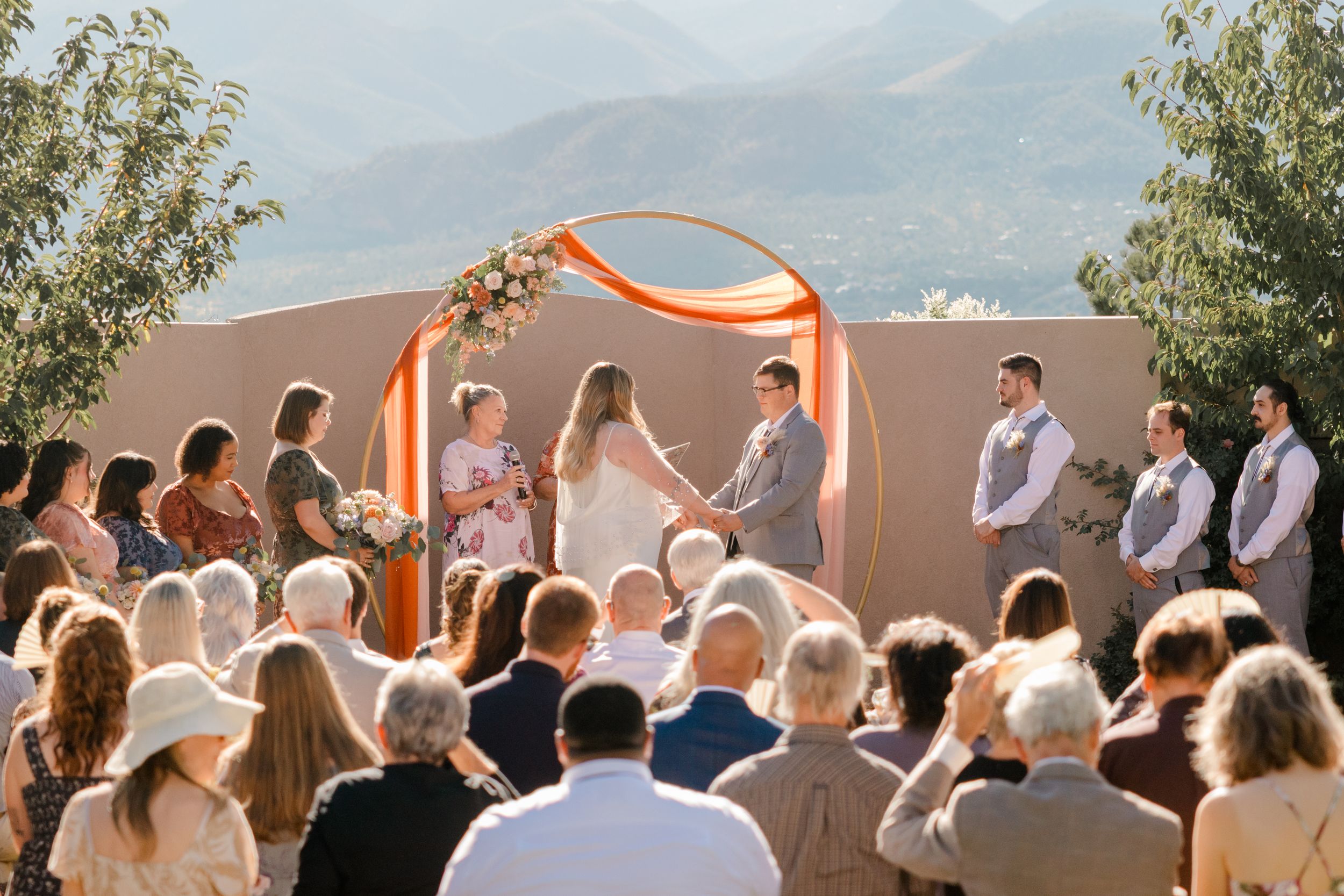 Outdoor wedding ceremony under a bright orange fabric arch with mountains and desert landscape in the background.