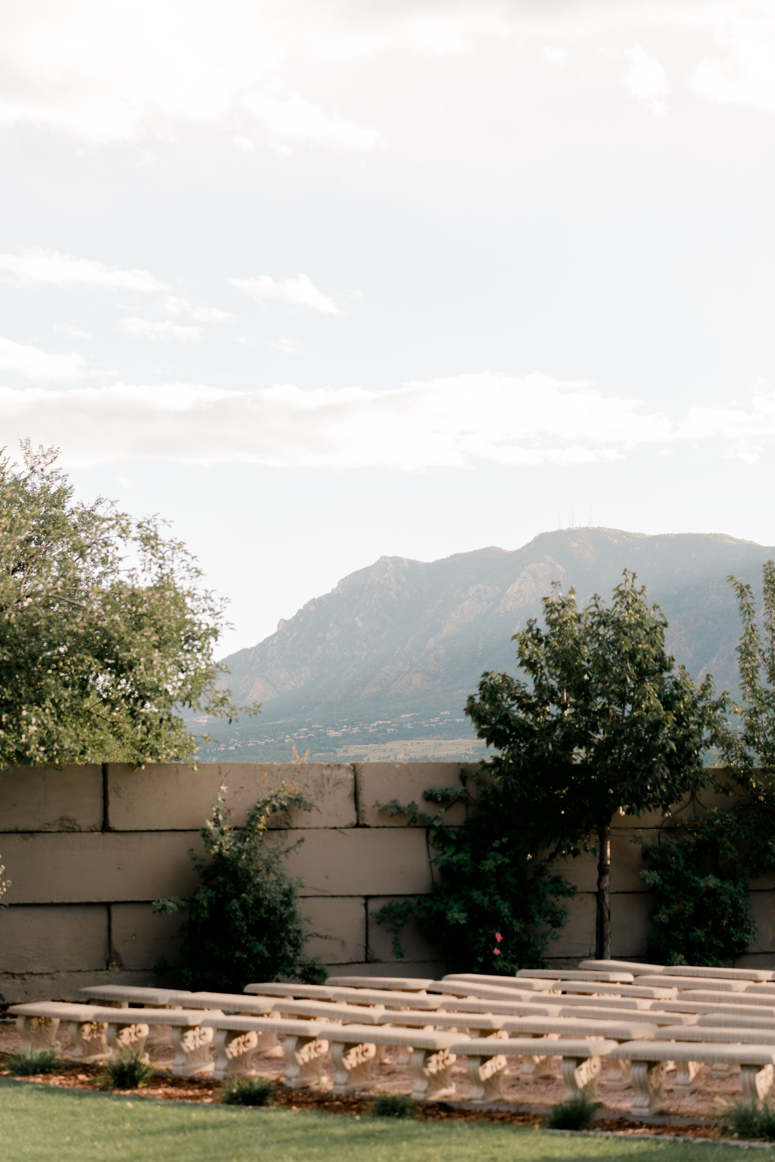 Stone benches arranged in rows facing mountains with adobe walls and trees in the background under a cloudy sky.