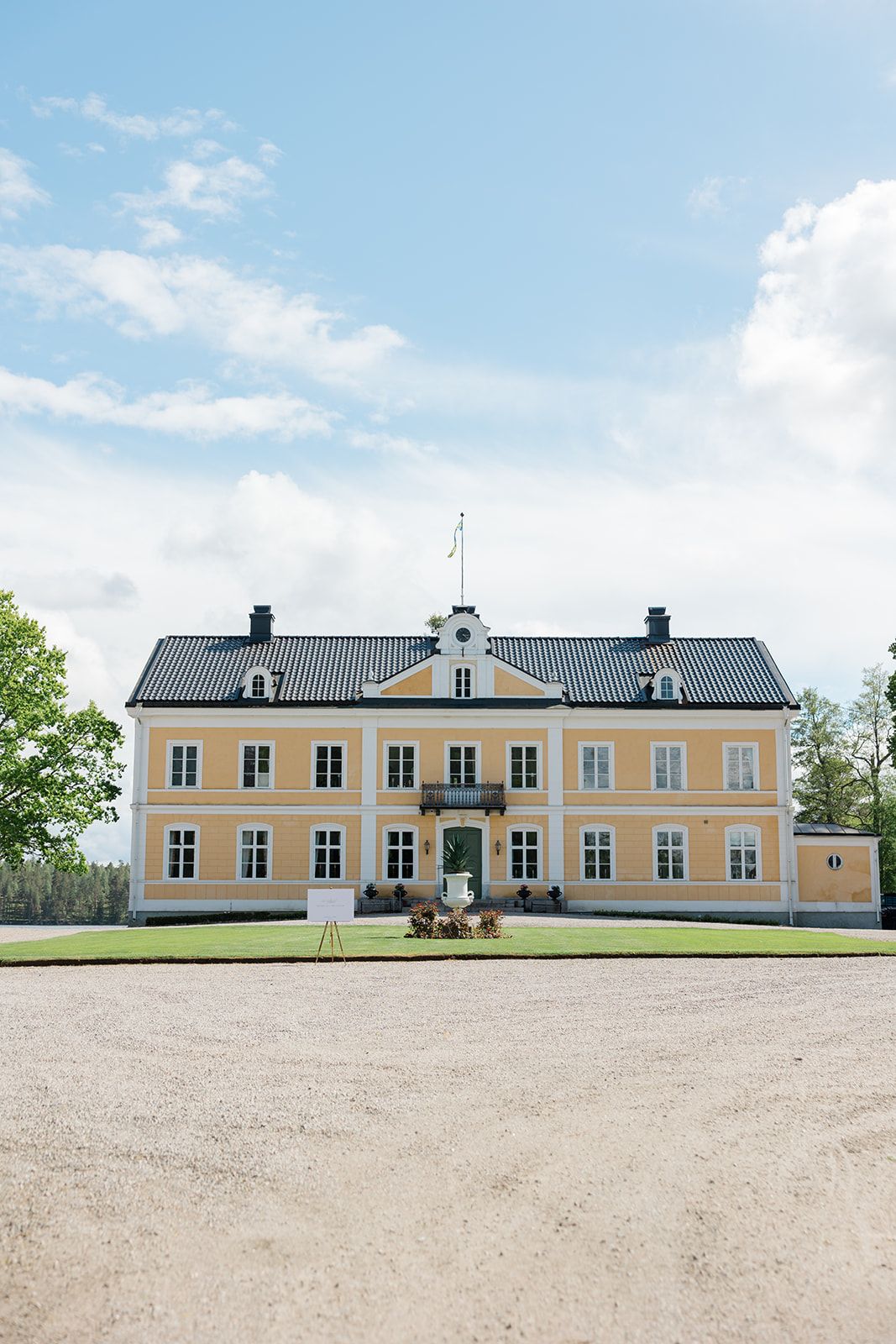 A long yellow manor house with black roof and white trim sits atop a gravel driveway against a blue summer sky.