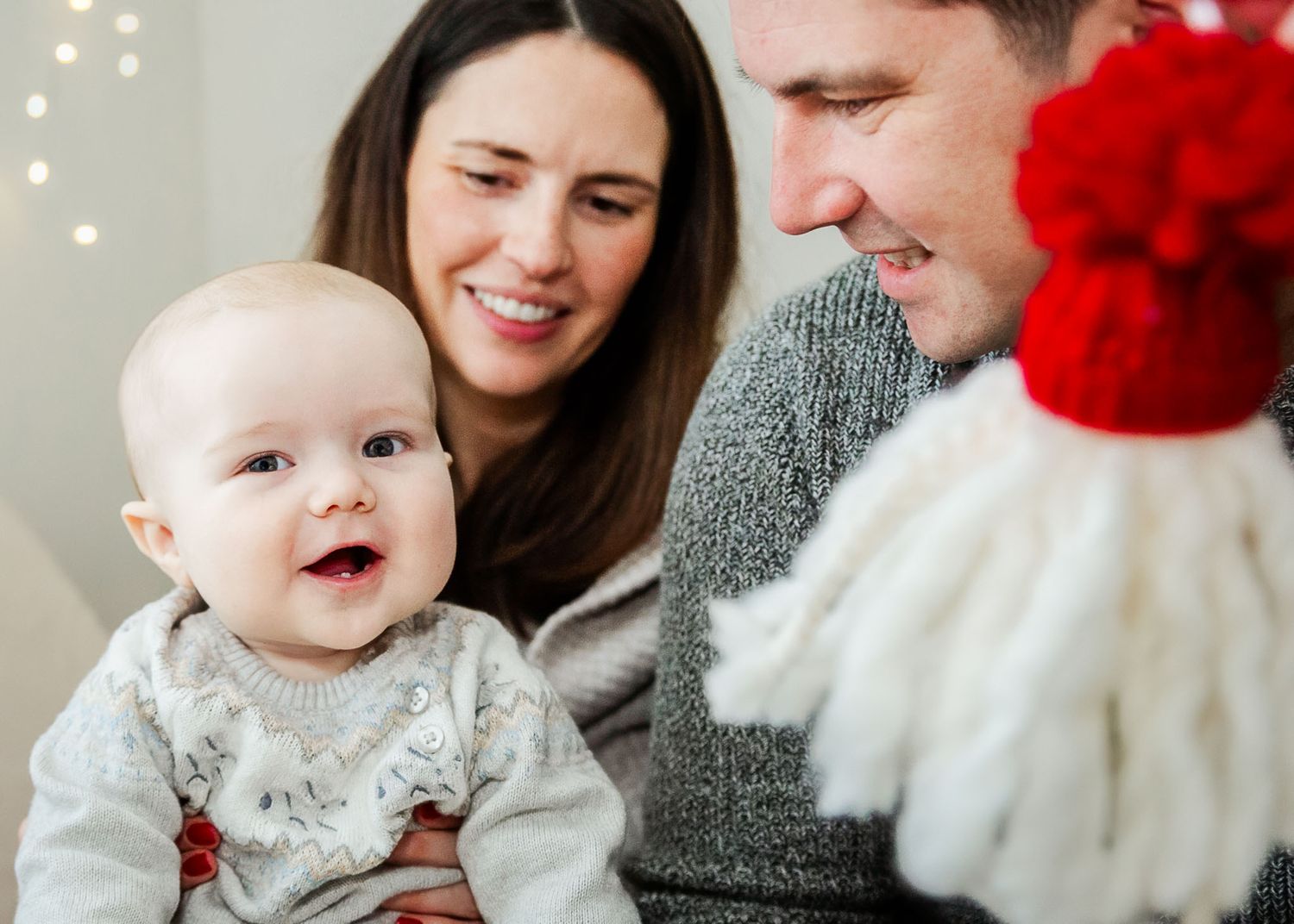 A baby smiles and looks to camera as he sits on his mum's knee and his dad holds up a gonk. Natural in-home Christmas photoshoot in Bristol