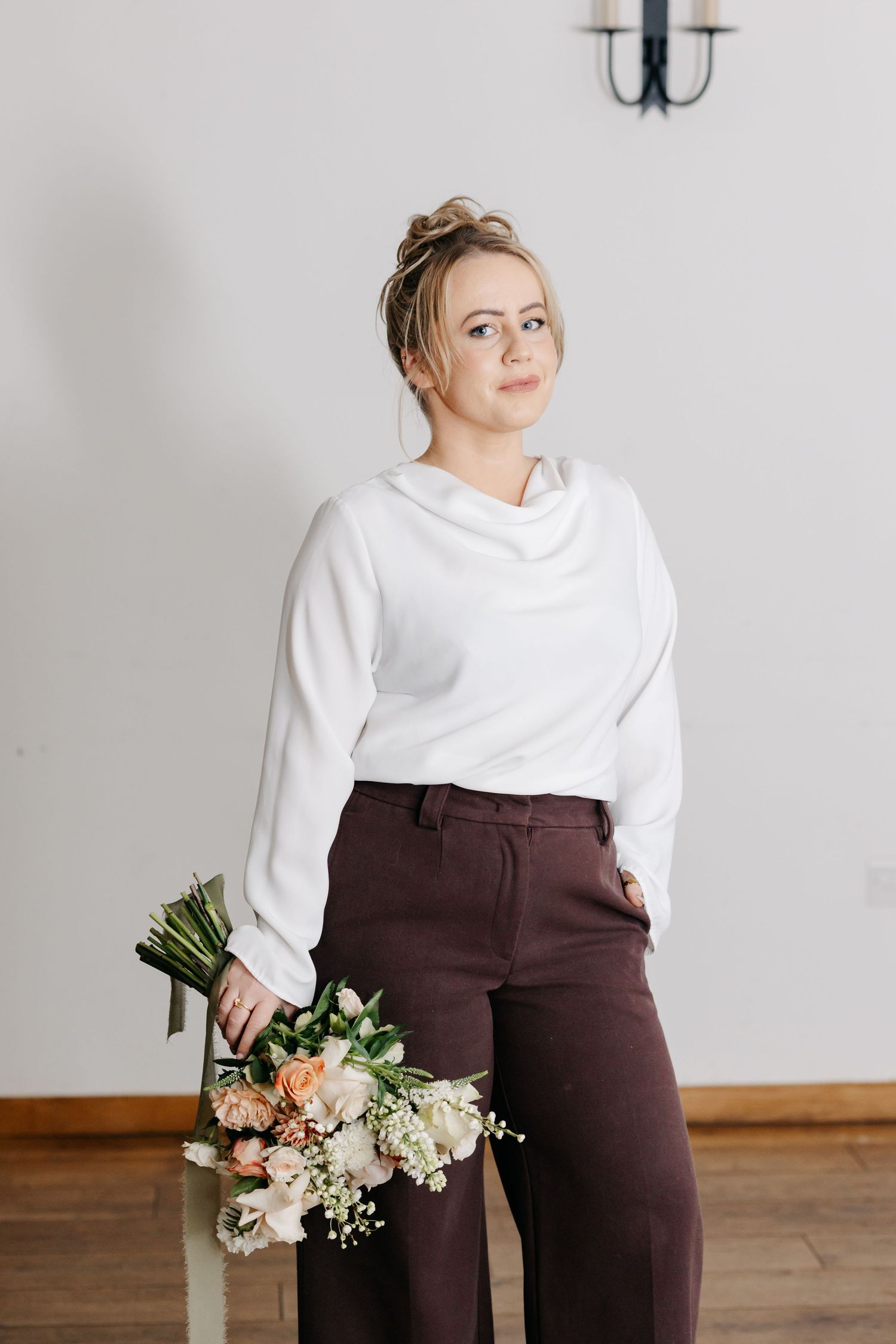 A person in a white sweater and brown pants poses elegantly with floral arrangements in a minimalist studio setting.