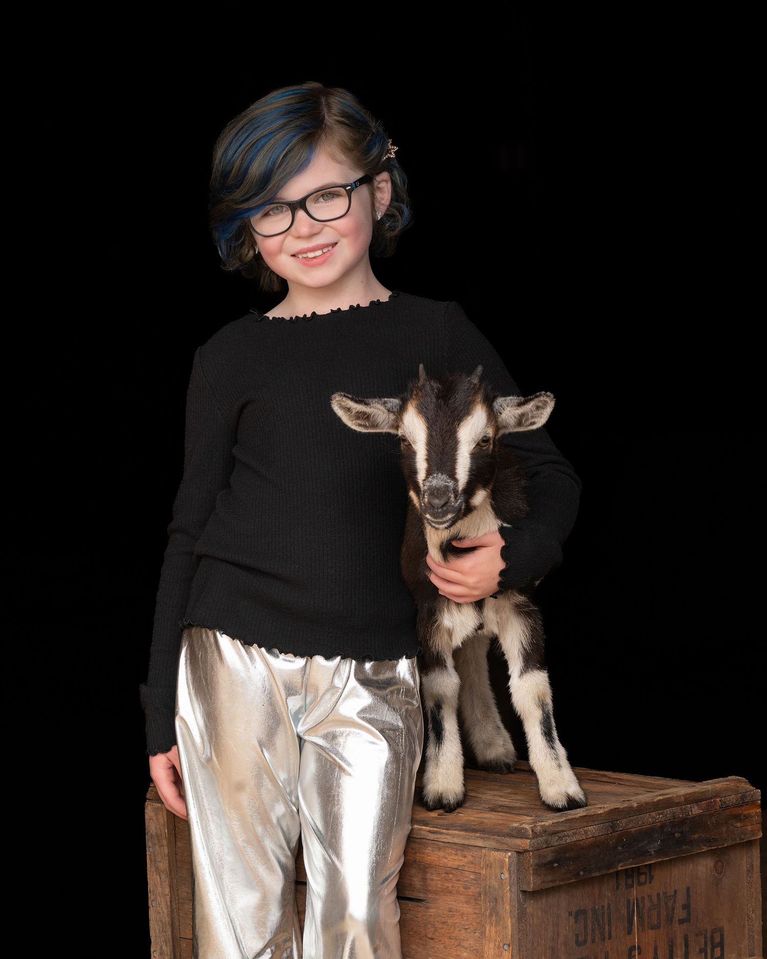 A small husky puppy sits on a wooden crate against a black background while someone in dark clothing looks on.