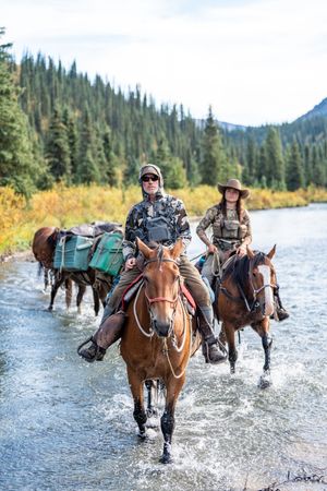 Riders crossing a mountain stream on horseback with pack horses in tow.