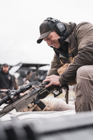 Person adjusting scope on tactical rifle while wearing protective gear and cap.