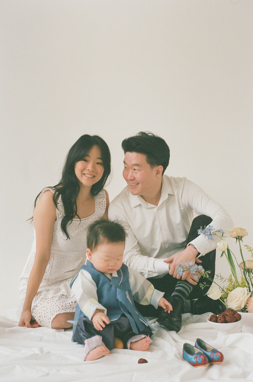 A family in white clothing shares tender moments together on a light-colored bed with white flowers and decor.