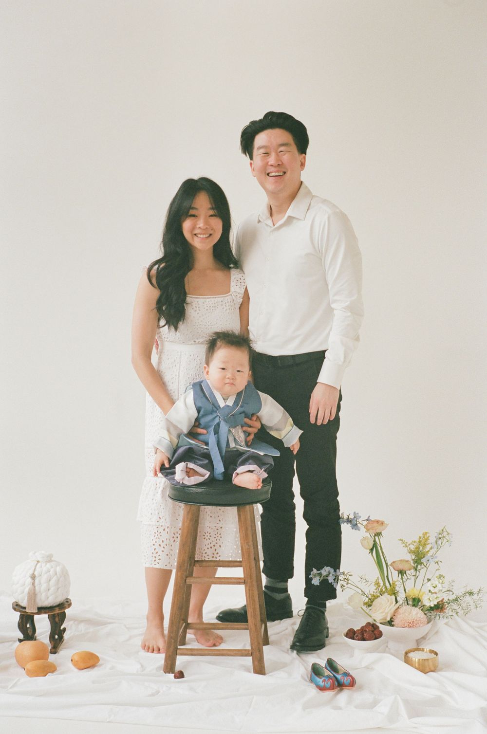 A minimalist family portrait series in white studio setting with wooden stool and floral arrangements.