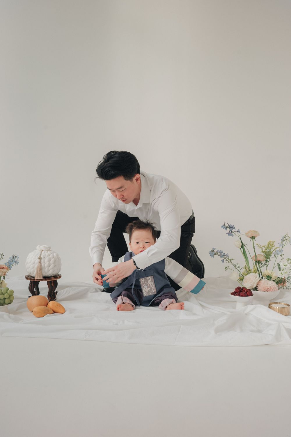 Parent and child sitting on white backdrop with flowers and decor for minimalist studio photo session.