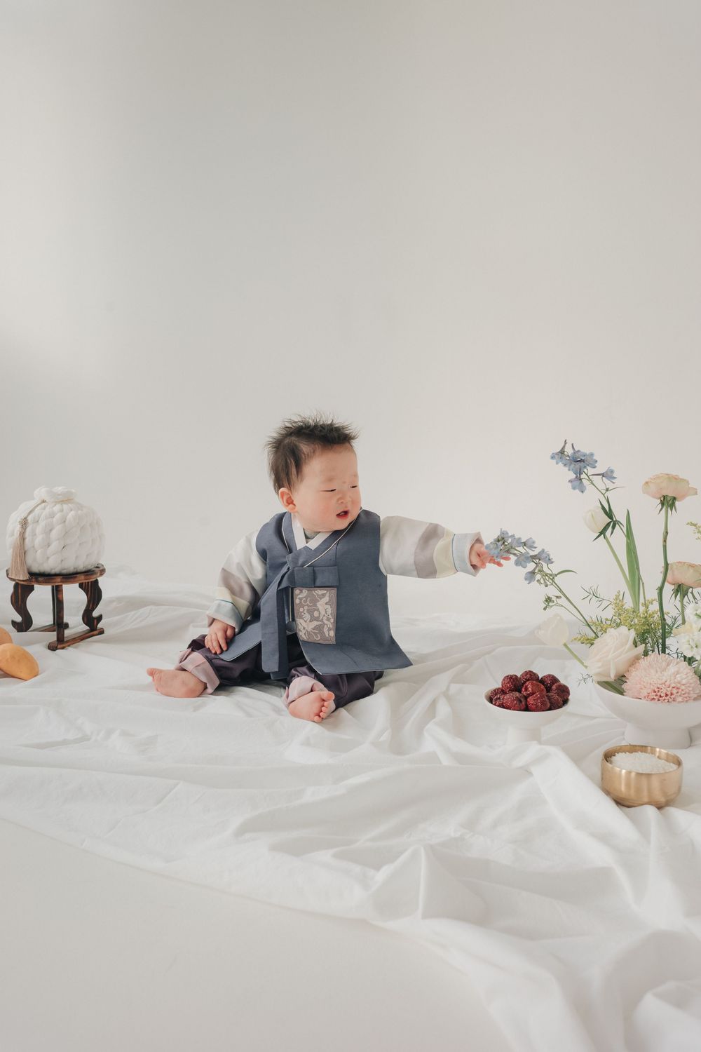 Baby in traditional Asian vest sits on white fabric reaching toward delicate flowers in minimalist studio setting.