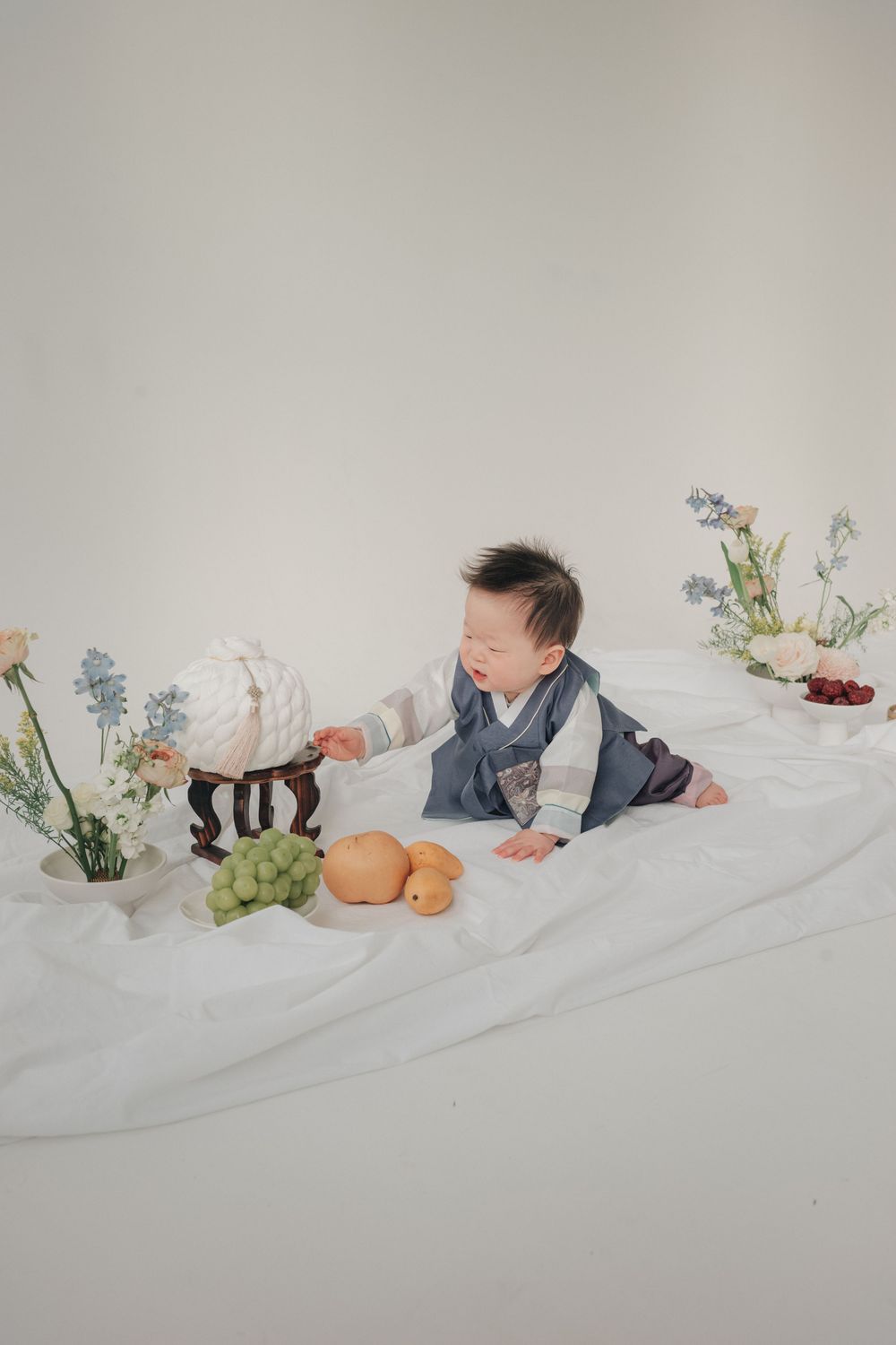 A minimalist photo series showing a baby lying on white sheets with delicate floral arrangements and decorative objects.
