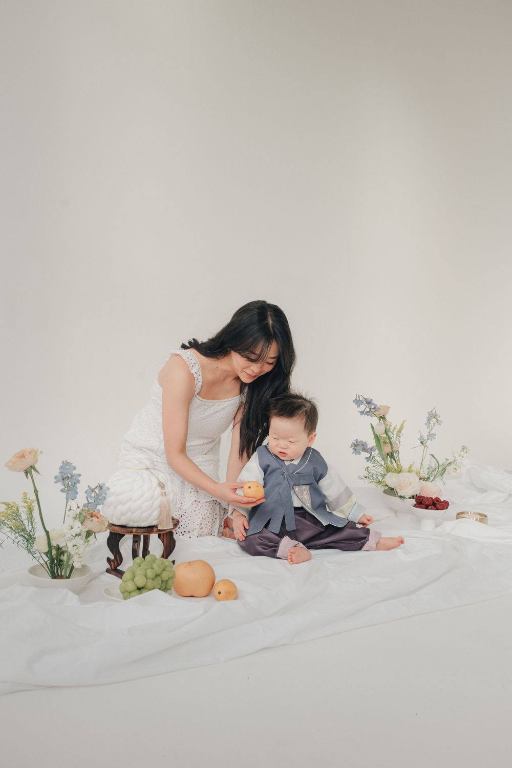 A parent and child in white clothing sit together on a light backdrop with flowers and oranges as props for a photo session.