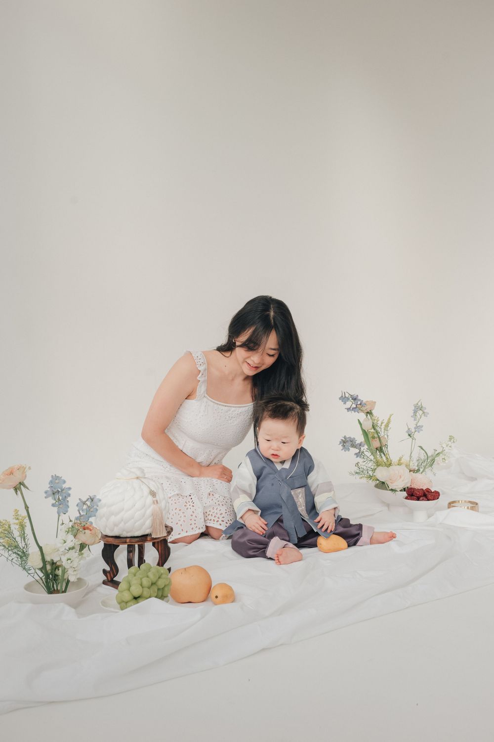 A mother and baby share tender moments on a white backdrop with delicate flowers arranged beside them.