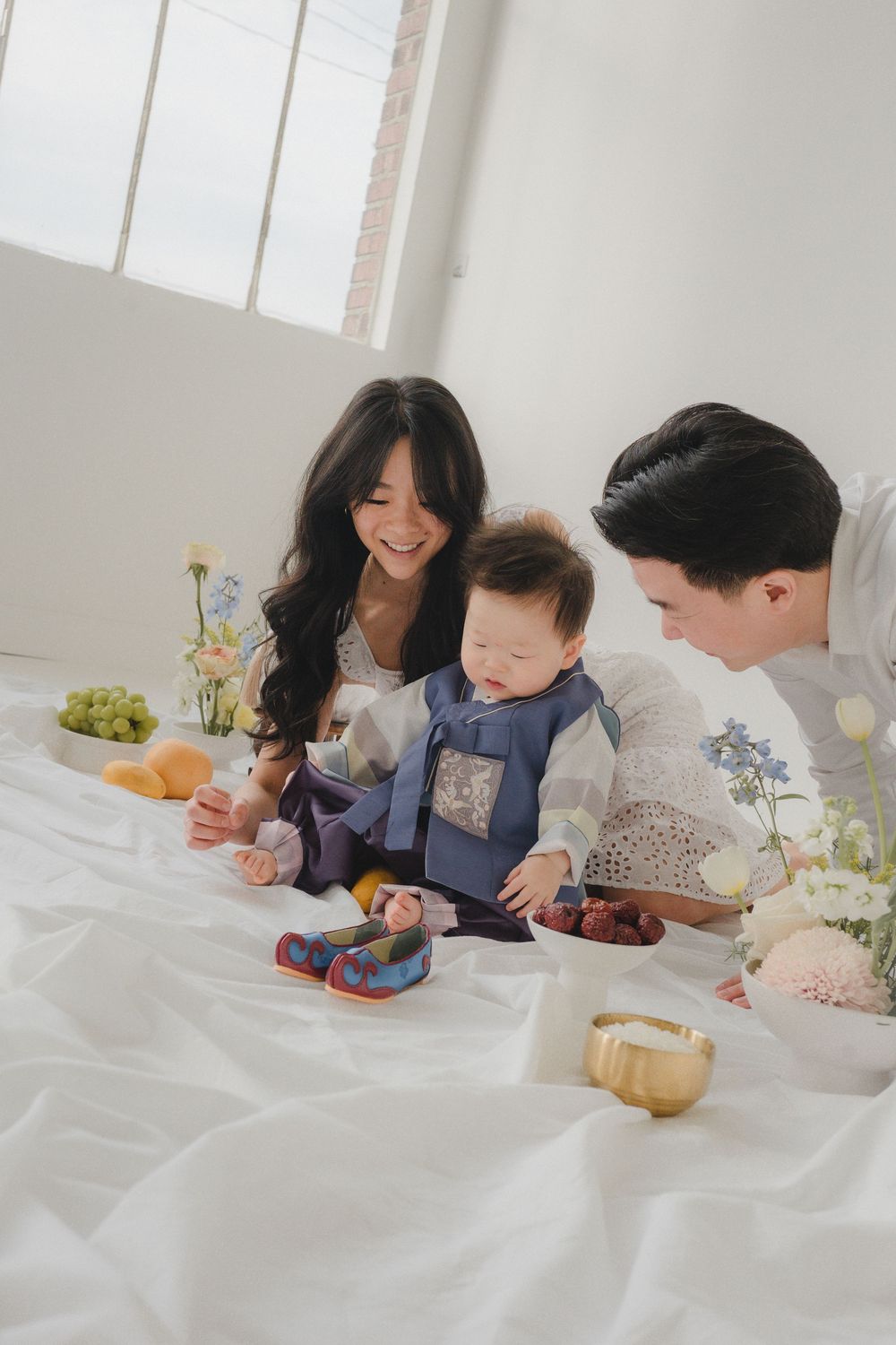 Family enjoying a cozy picnic indoors on white linens with fresh fruits and snacks spread out on the floor.