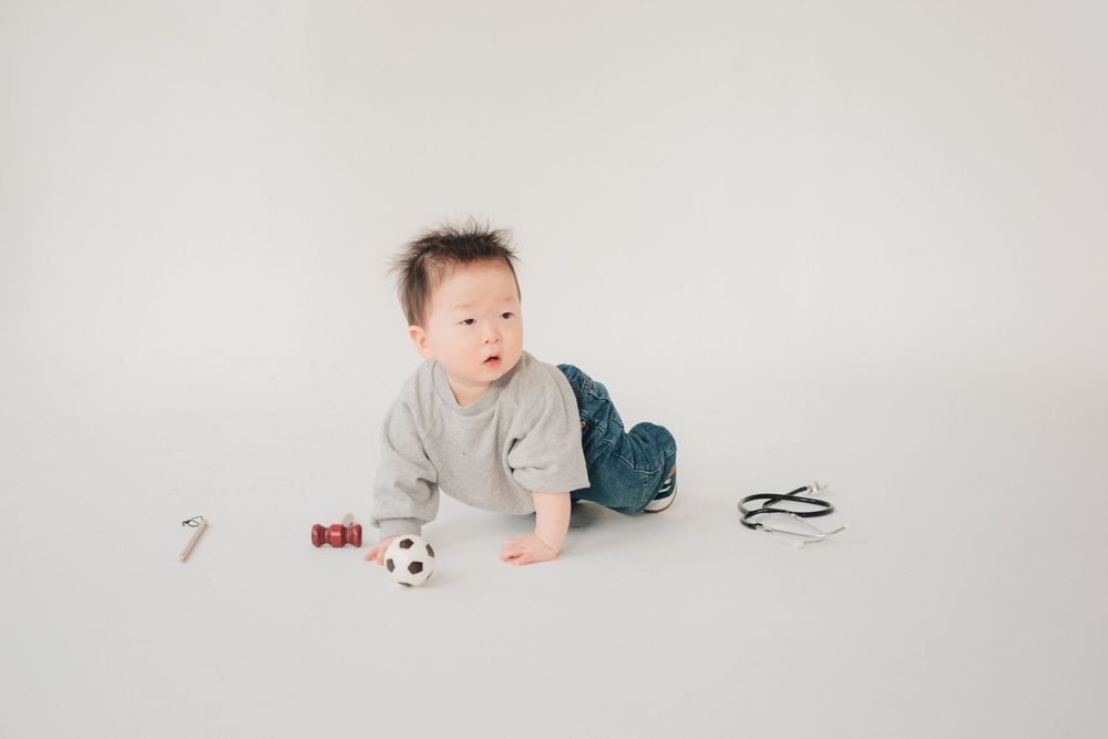 A baby in a gray onesie lies on their stomach on a white backdrop with a small soccer ball nearby.