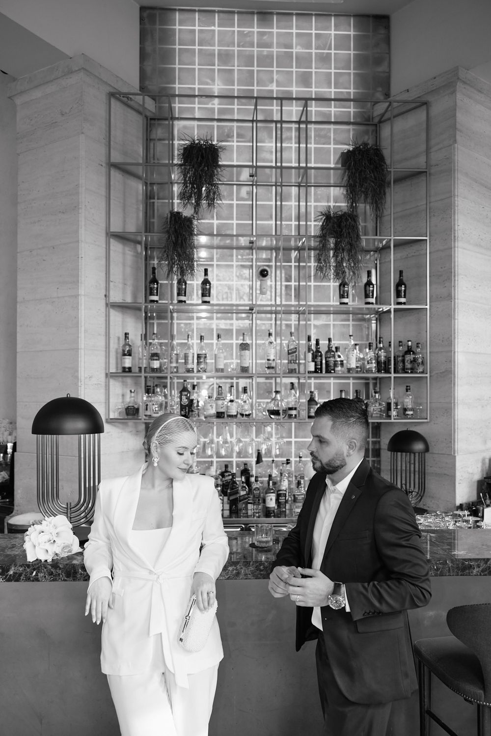 A couple in formal attire poses together in a series of black and white photos at a modern bar with glass shelving.