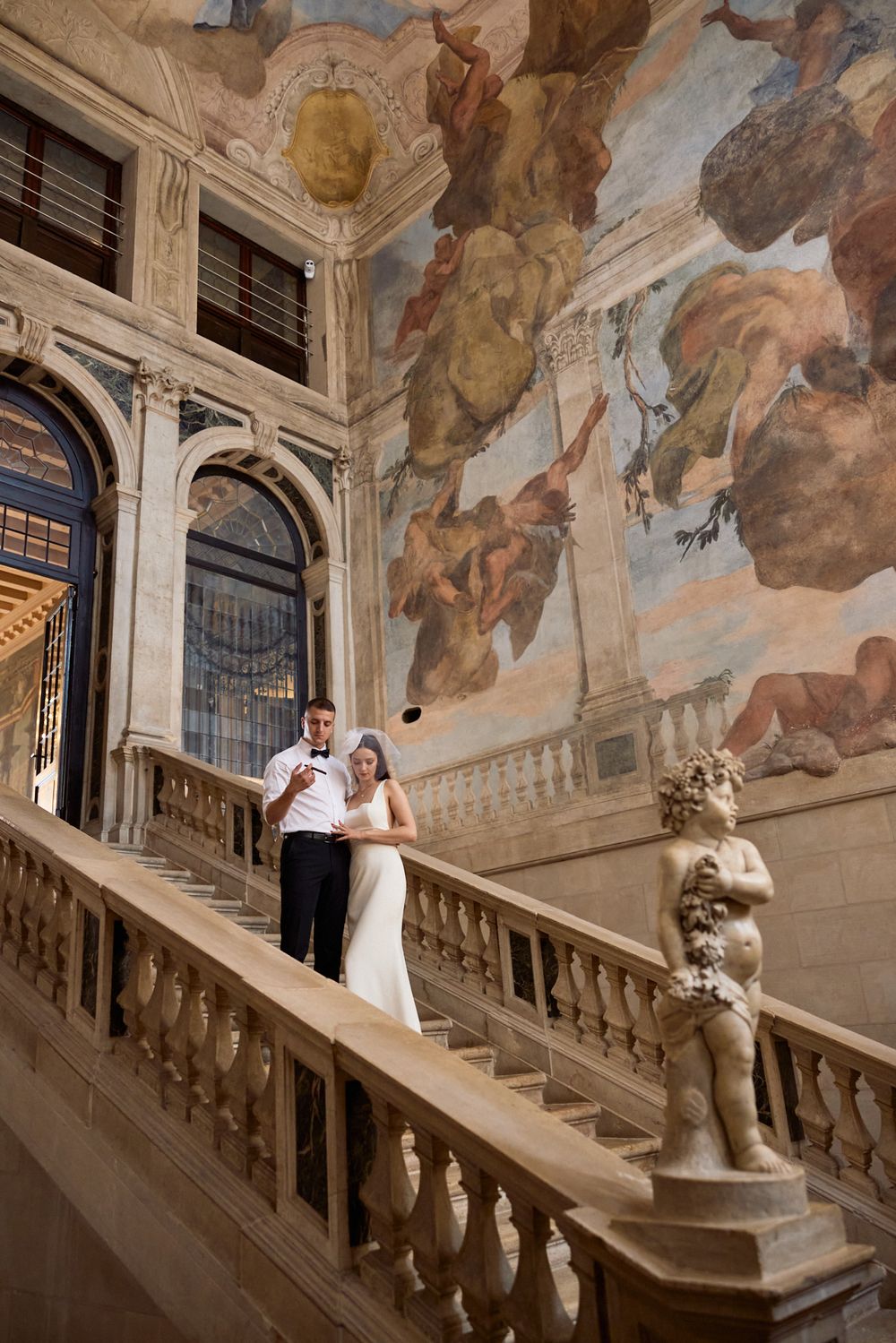 A wedding couple poses on an ornate baroque staircase with painted frescoes and sculptures in a historic palace.