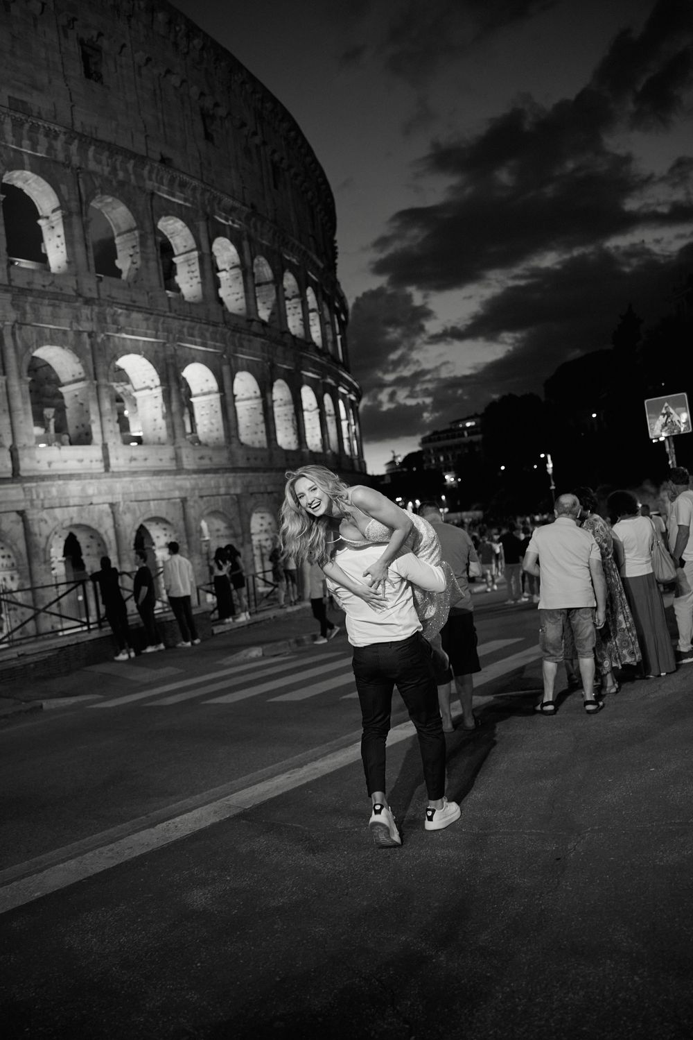 A black and white photo capturing a playful moment outside the illuminated Colosseum at dusk.