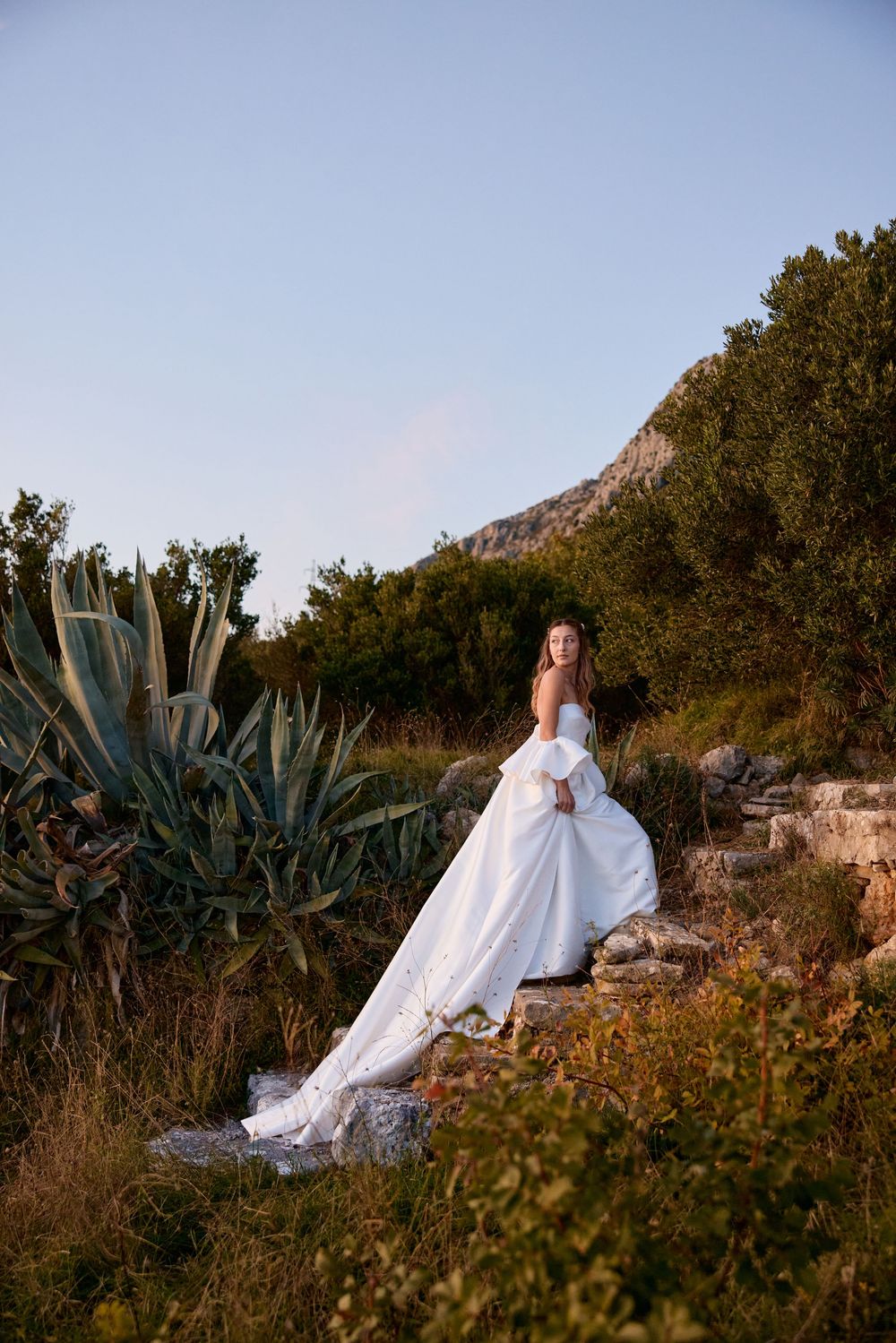 A white wedding dress flows elegantly against a rugged Mediterranean landscape with agave plants at sunset.
