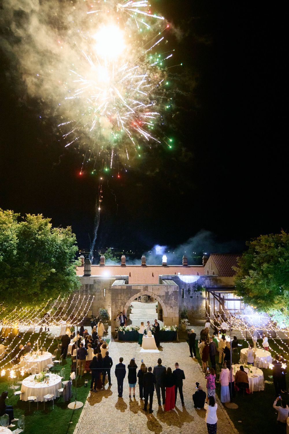 Wedding guests gather under string lights to watch a spectacular fireworks display during an outdoor evening celebration.