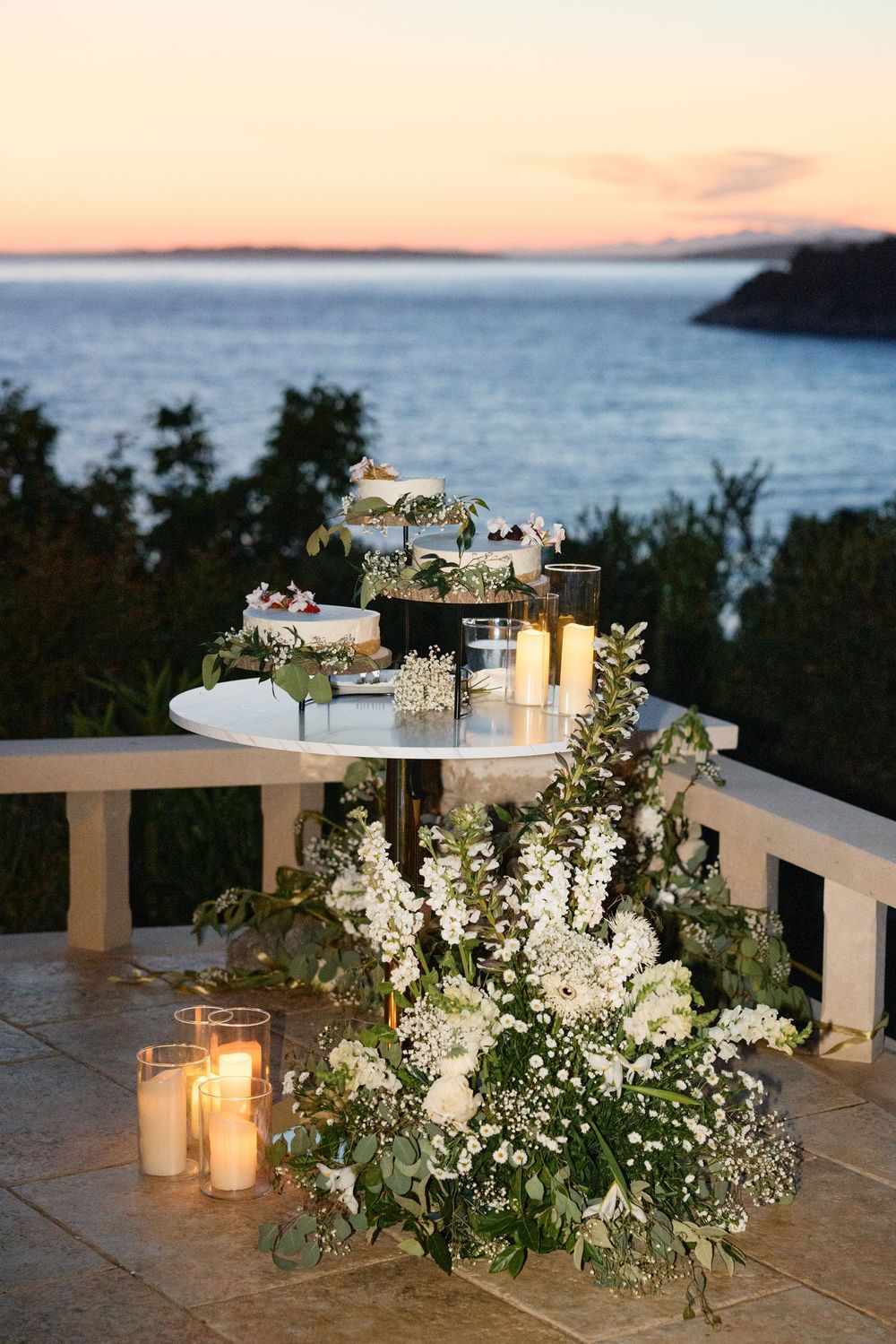 White floral arrangements and glowing candles line an oceanfront terrace railing at sunset.