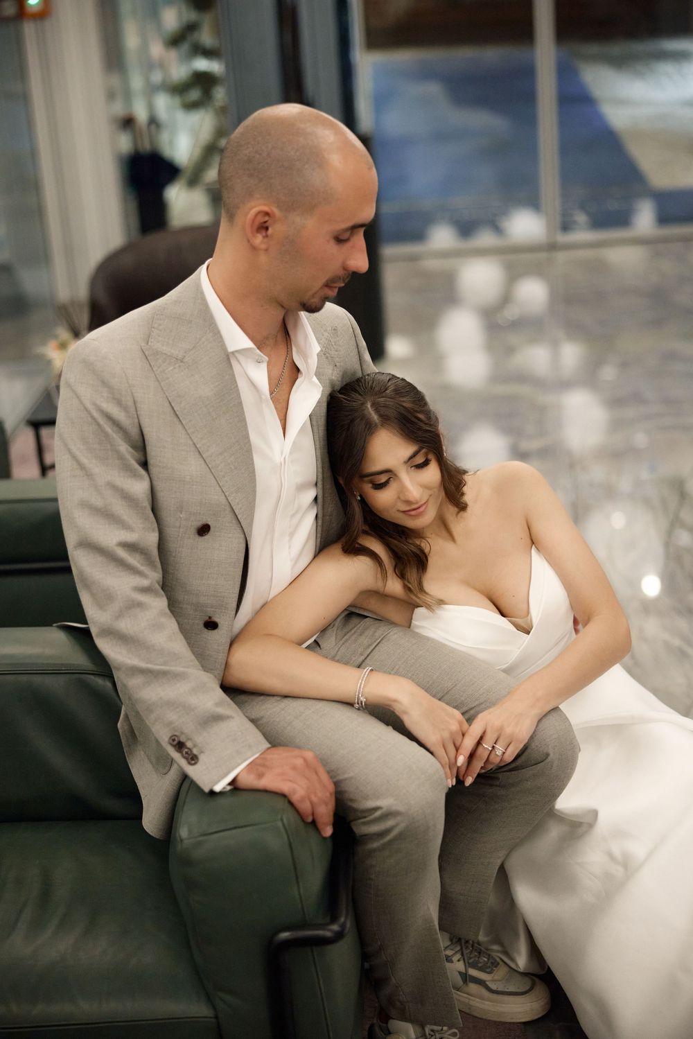 A couple shares an intimate moment on a dark green couch, with the bride wearing a white gown and groom in a gray suit.