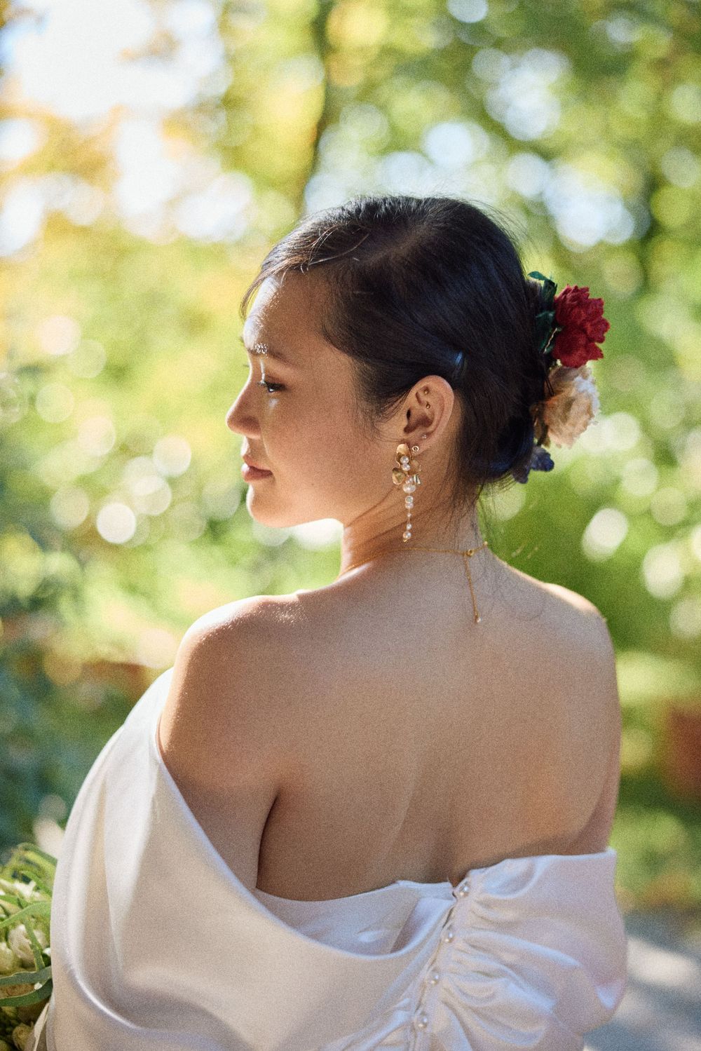 A bride in a white off-shoulder dress poses outdoors with red flowers in her hair and elegant dangling earrings.