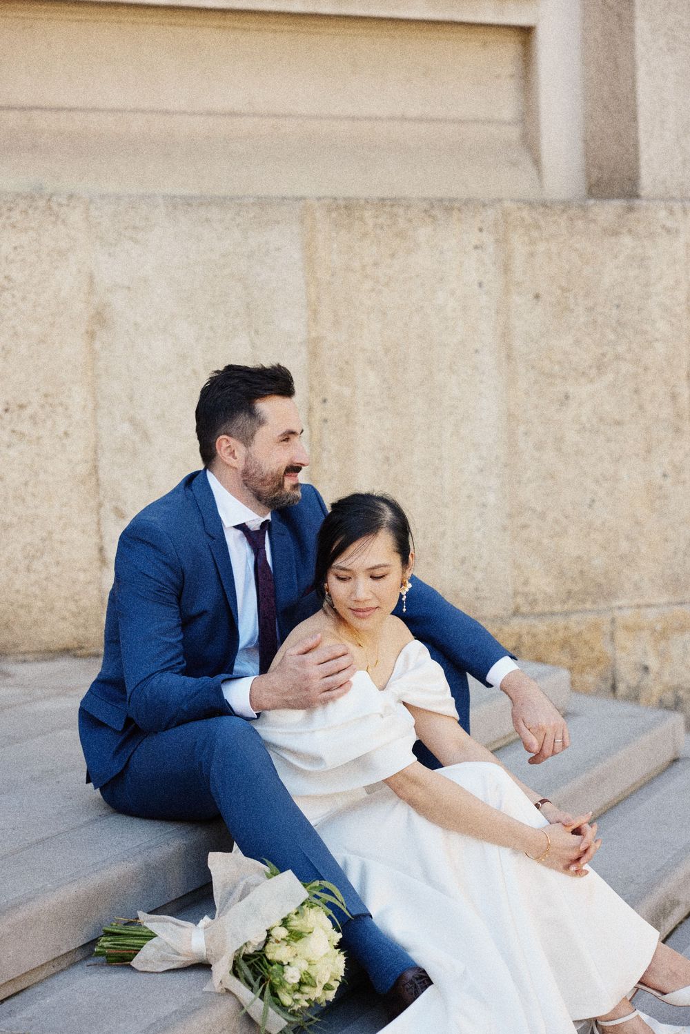 A couple in wedding attire sits on stone steps sharing an intimate moment in a romantic outdoor setting.