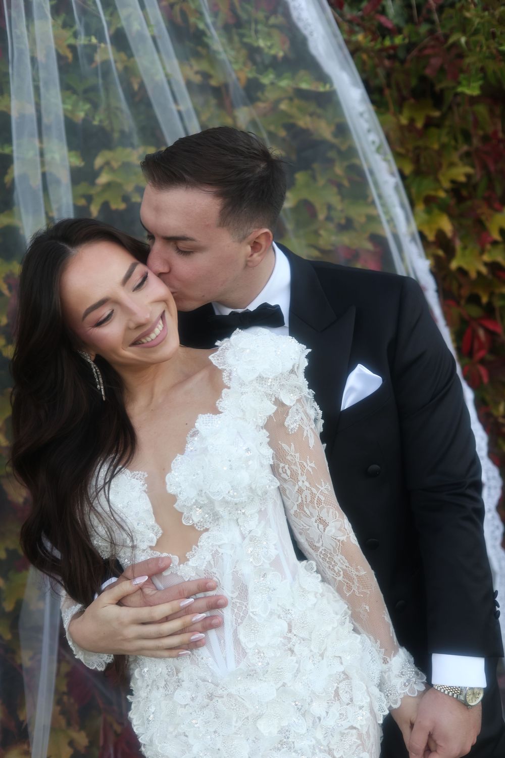 Groom in black tuxedo kissing bride in white lace dress, both smiling, with autumn foliage backdrop.