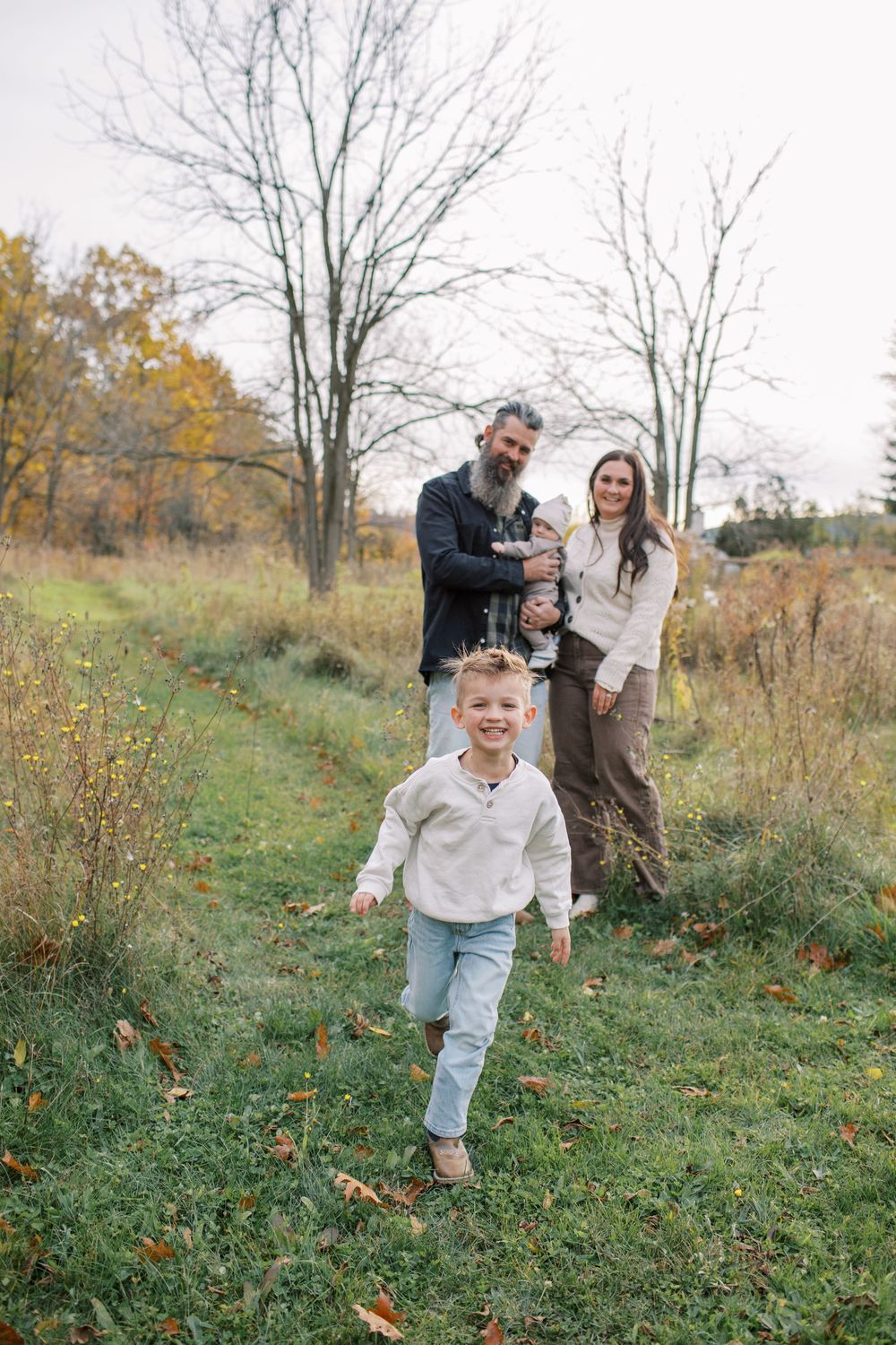 A family playfully chases each other through a grassy autumn field on a cloudy day.
