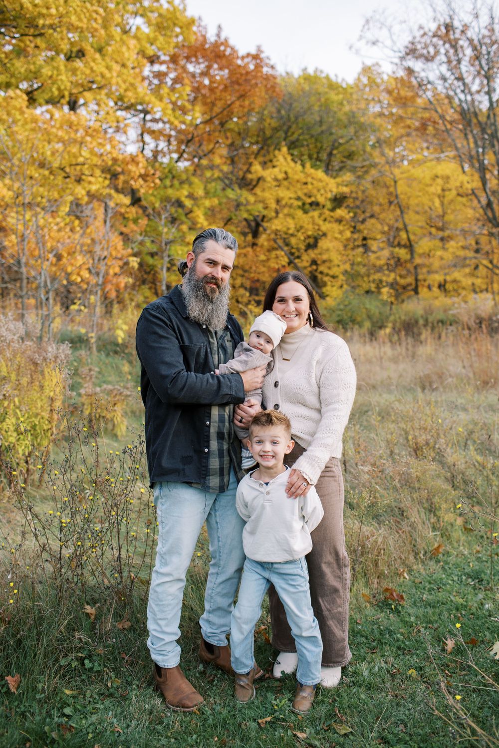 A family enjoys a fall photoshoot together in a field with golden autumn foliage in the background.