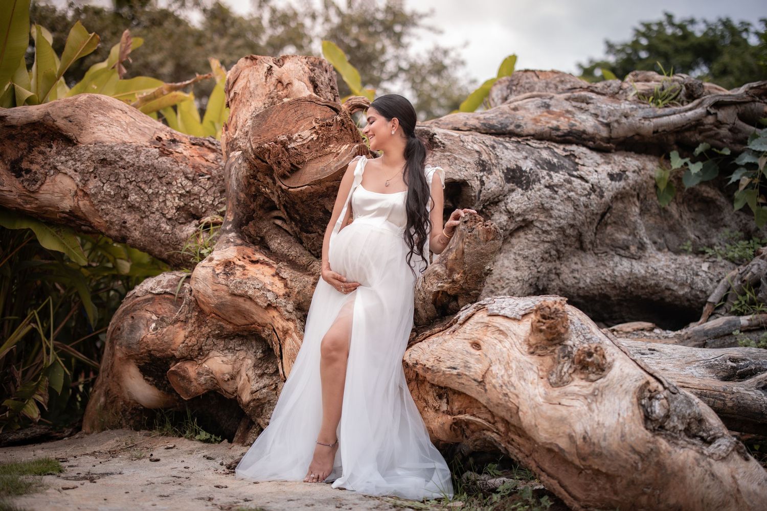 A couple in white dresses poses on driftwood at a romantic beach photoshoot during sunset.