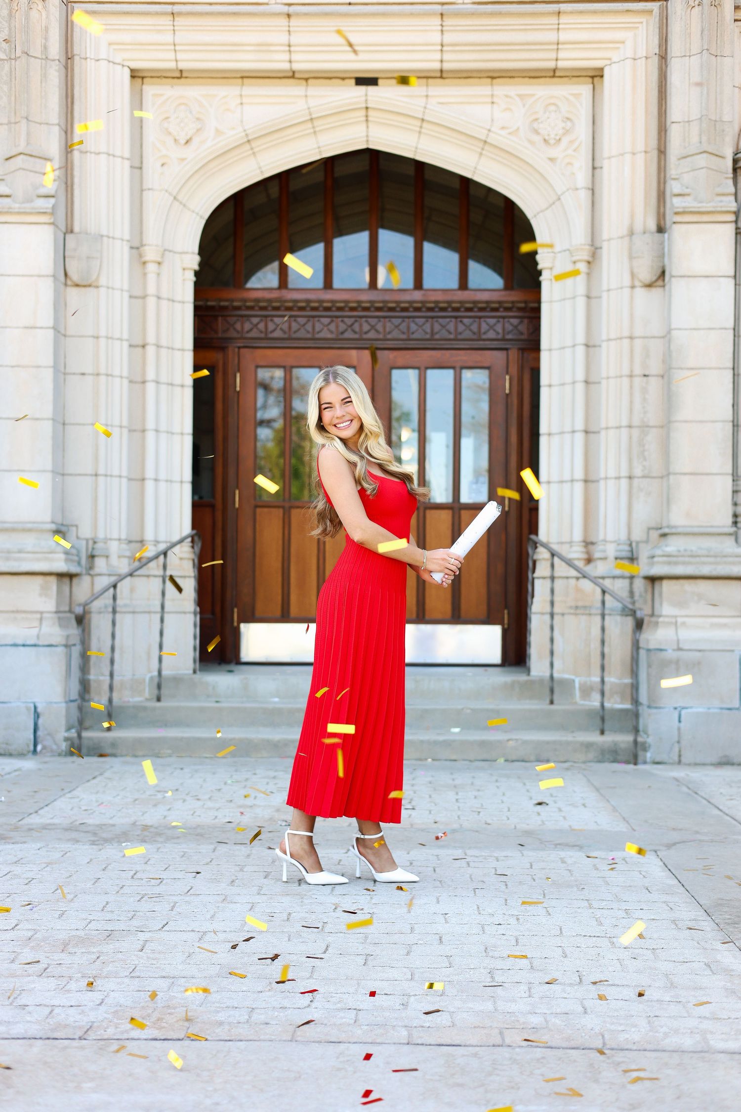 Person in red dress twirls joyfully under falling autumn leaves in front of ornate stone archway entrance.