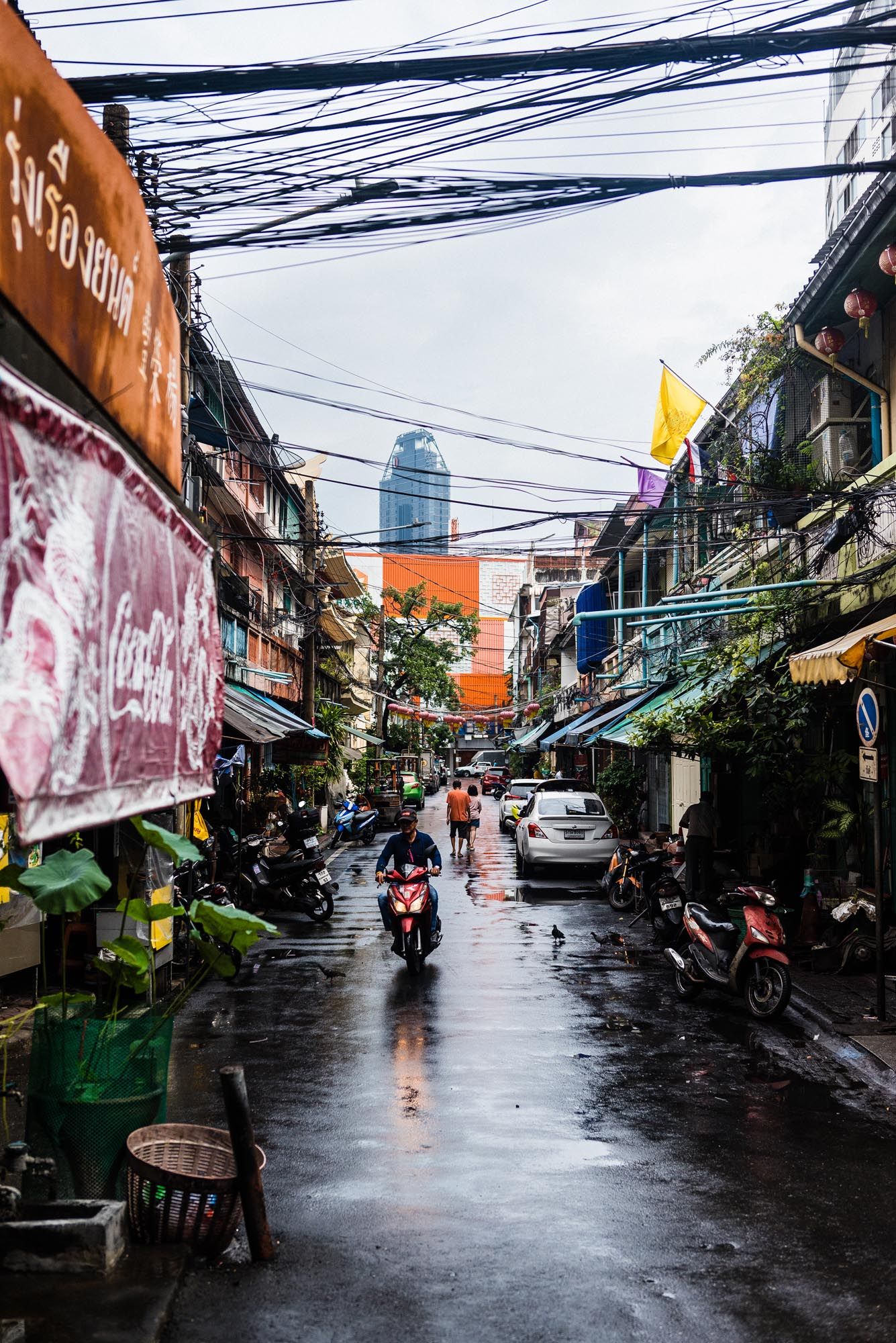Rainy street scene in Bangkok with tangled power lines overhead and motorcycles parked along narrow alley.