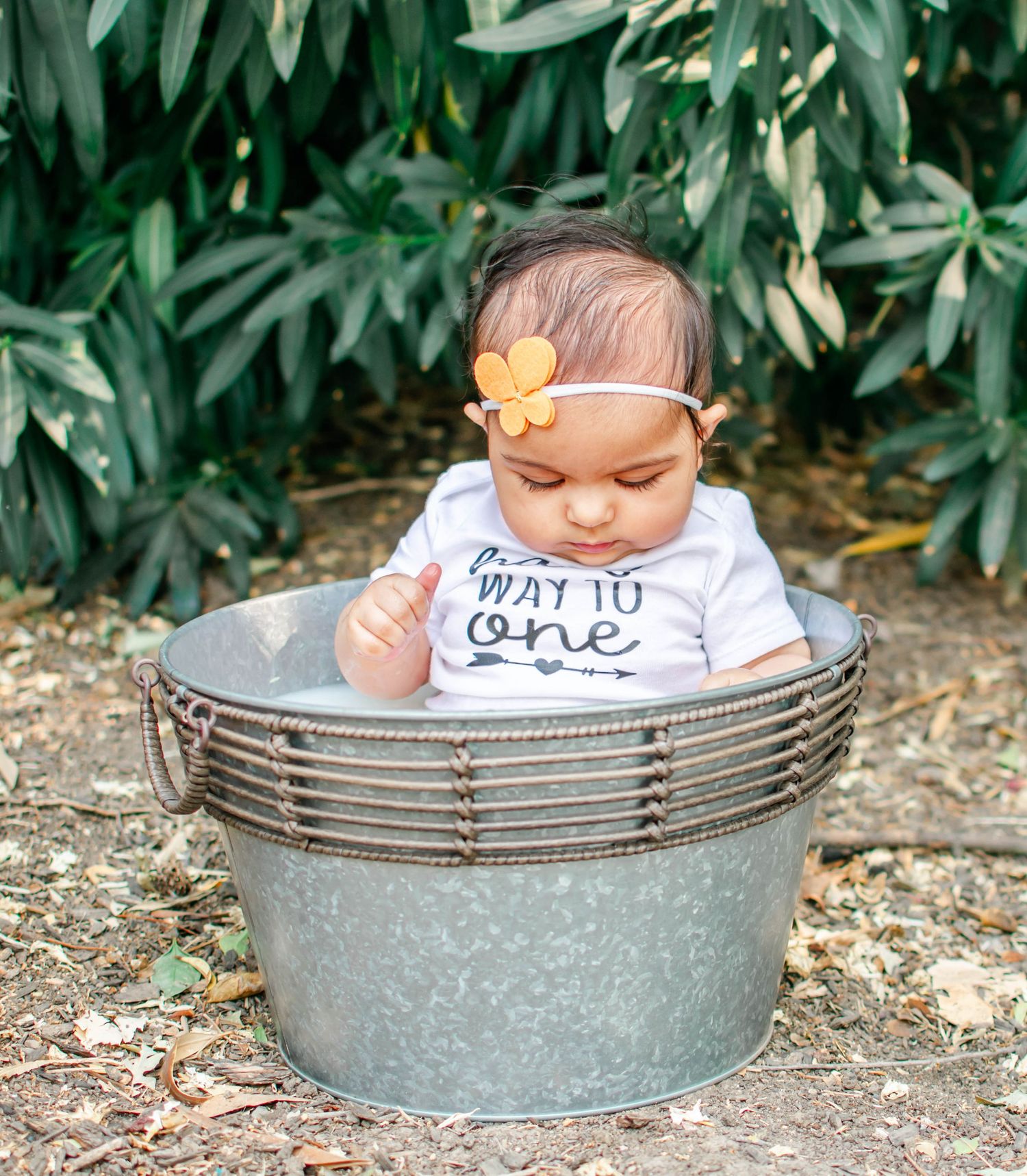 Small child wearing a 'Way to One' shirt sits in a rustic metal washtub outdoors near green foliage.