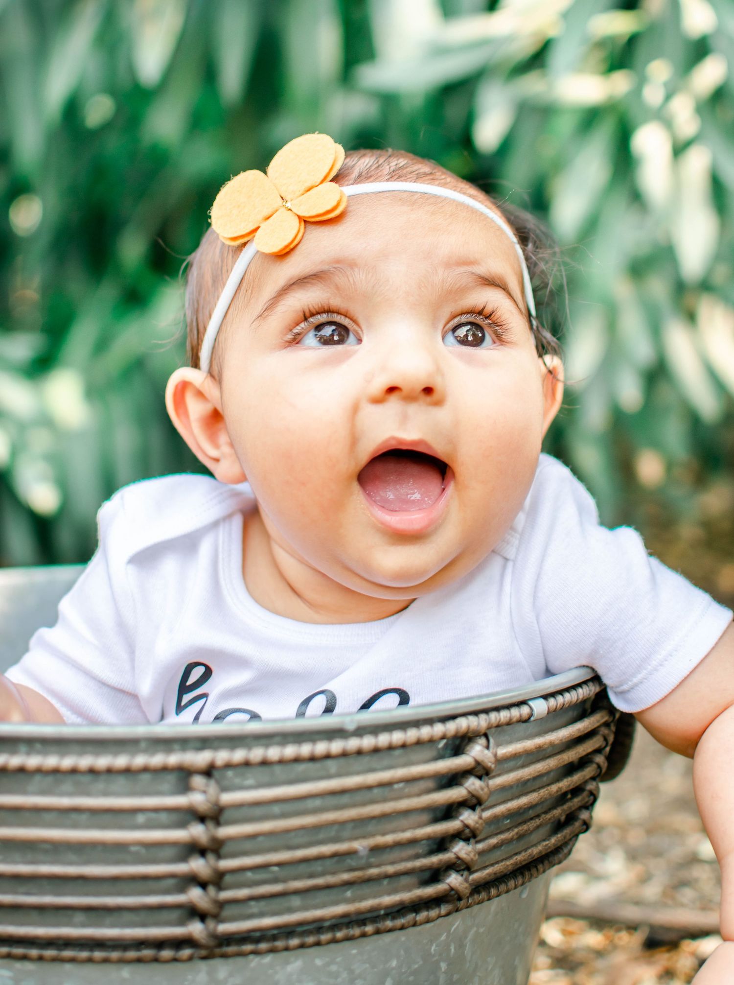 A baby wearing a white onesie and orange flower headband smiles while sitting in a wicker chair outdoors.