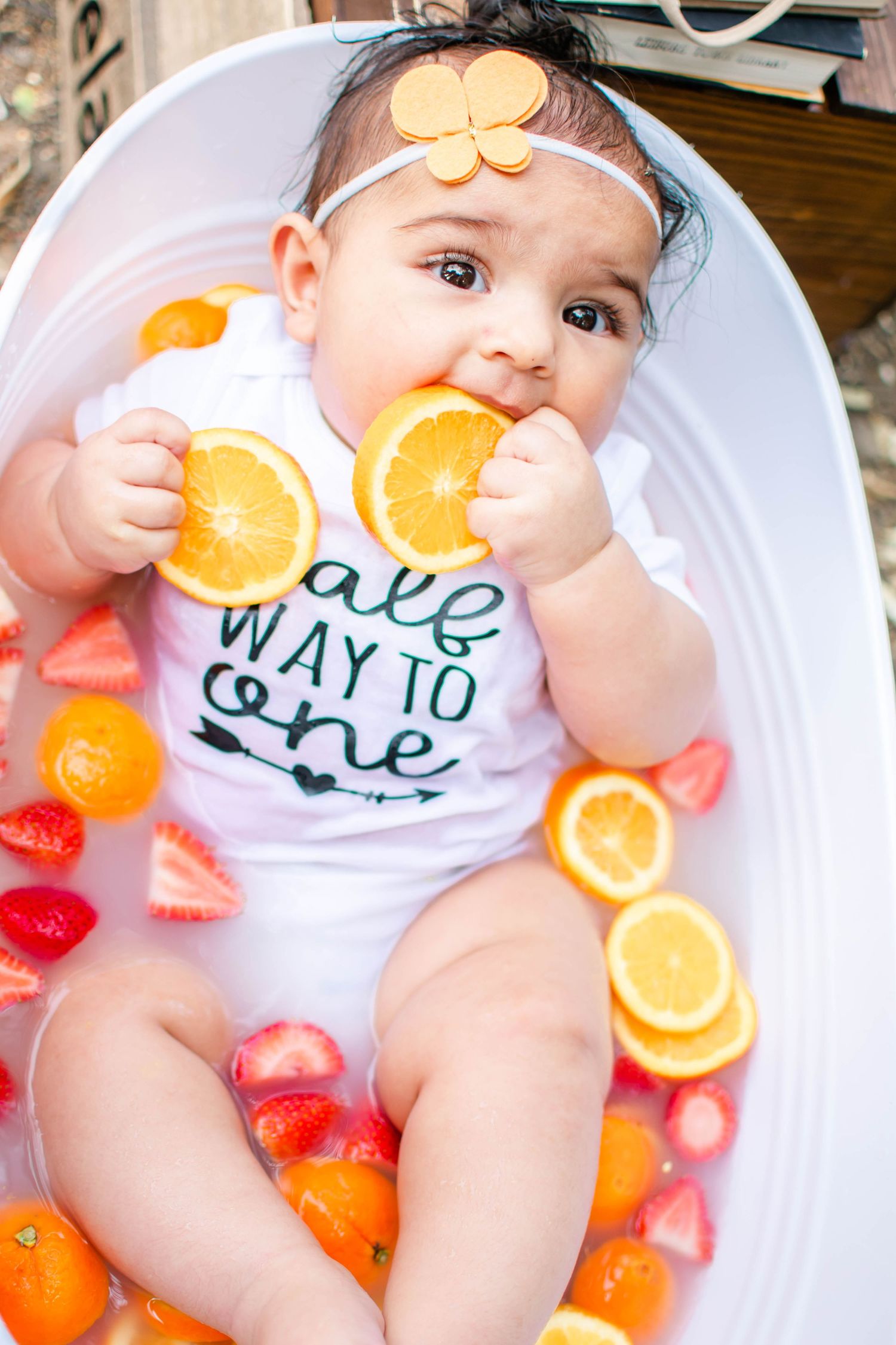 Baby in white onesie lies in bathtub surrounded by fresh oranges and red apples for creative milestone photo shoot.