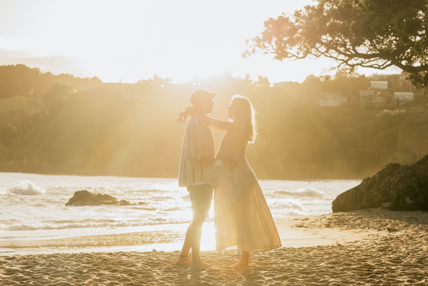 A couple in white and pink attire poses joyfully in a green outdoor setting on a cloudy day.