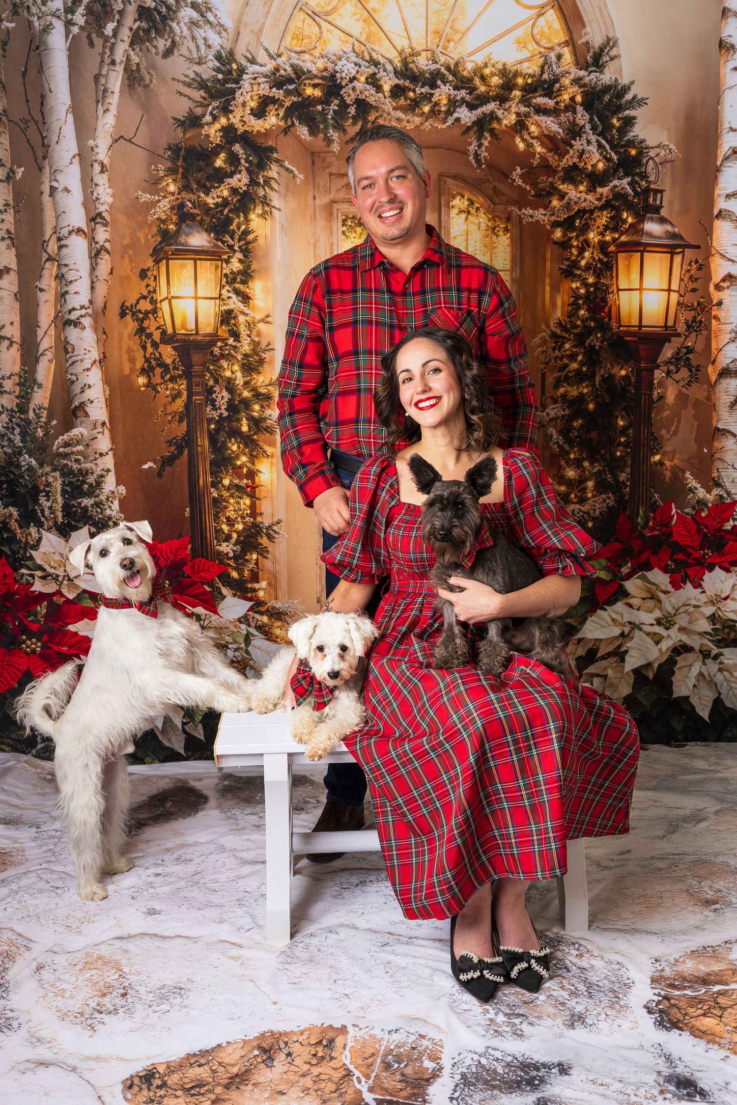 A couple in matching red plaid outfits poses with their white dog in a winter wonderland holiday setting with birch trees.