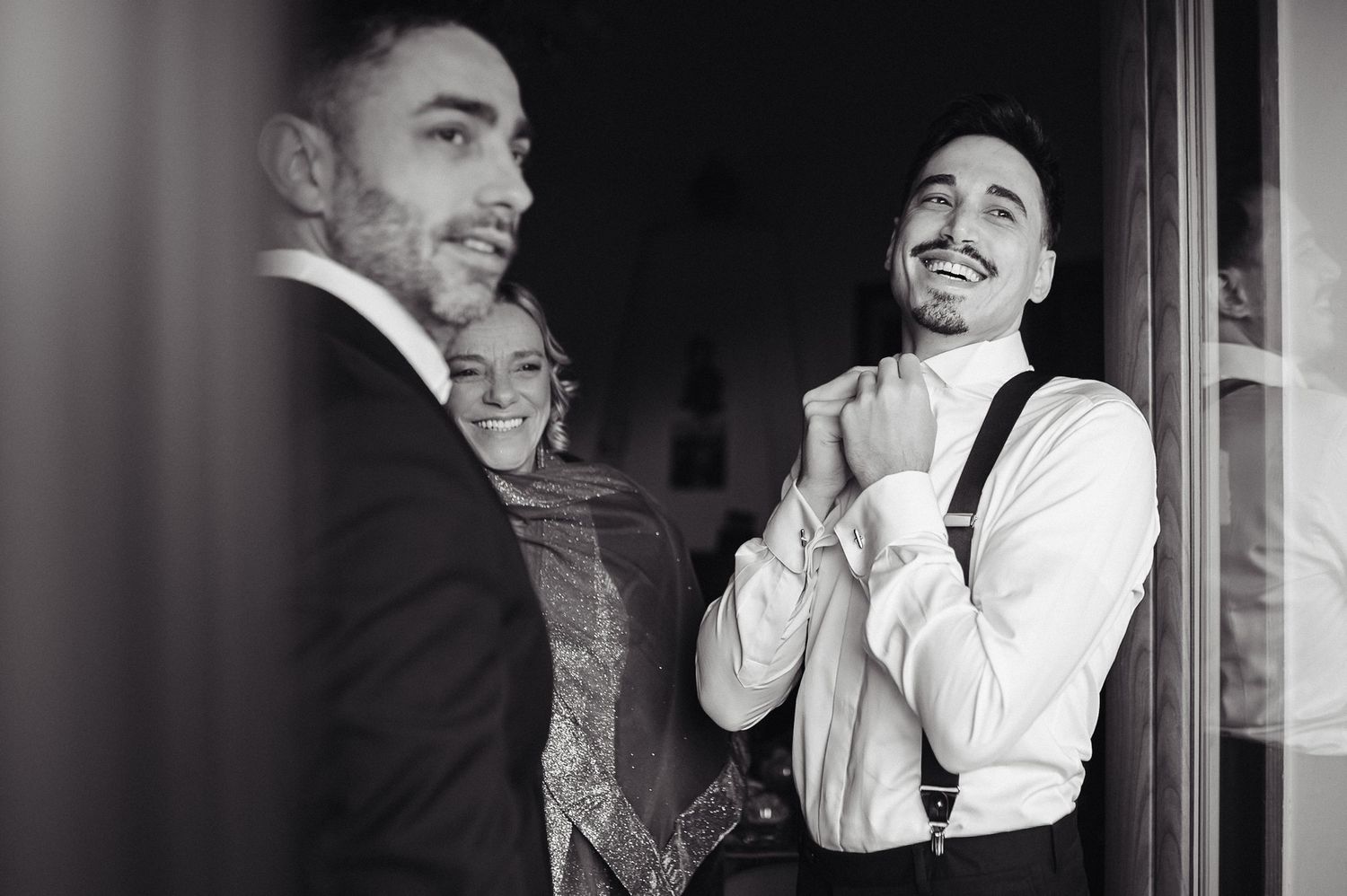 Foto in bianco e nero dello sposo sorridente con le bretelle, mentre i familiari lo osservano durante i preparativi del matrimonio.