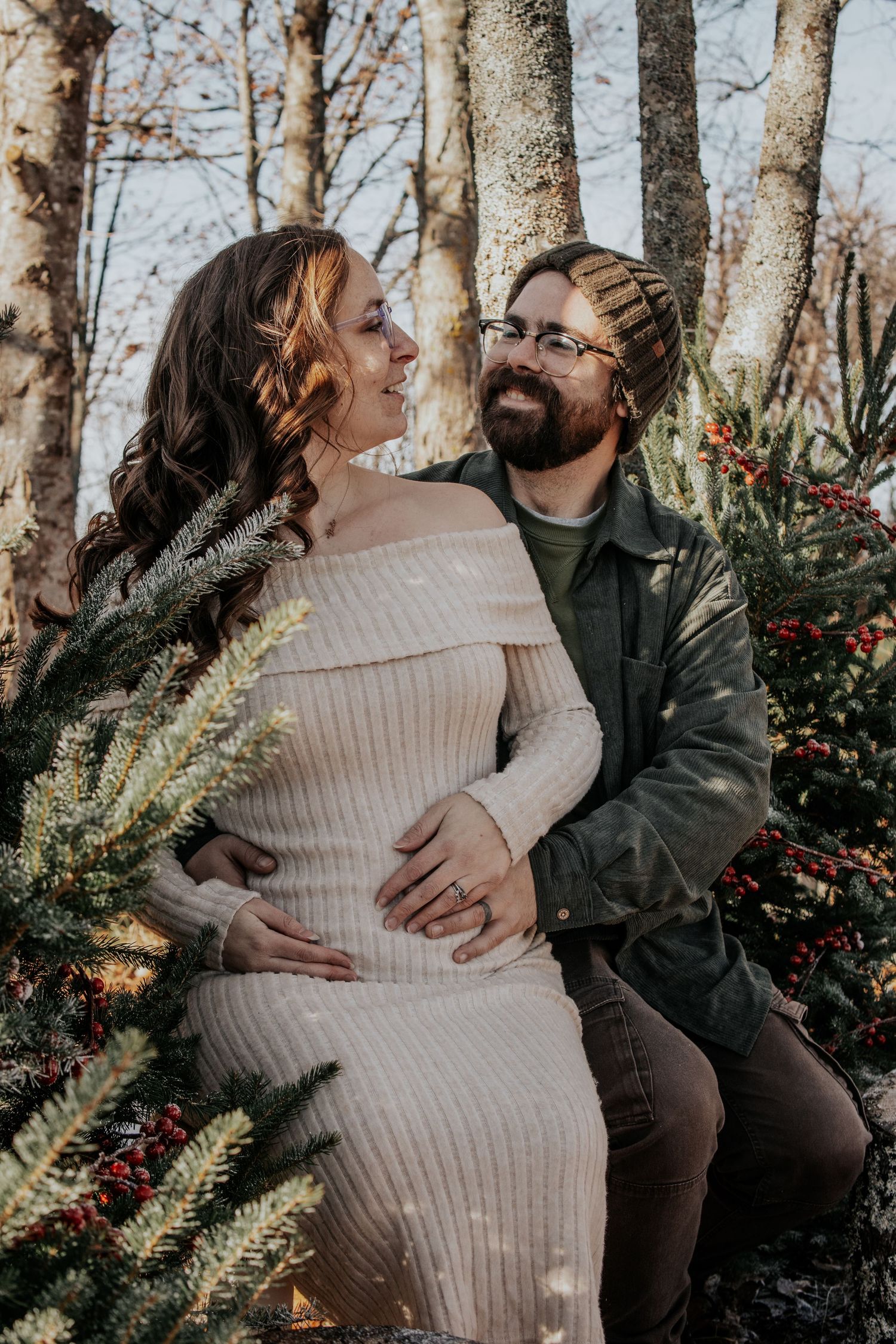 Expecting couple shares intimate moment outdoors among winter trees and red berries at sunset.